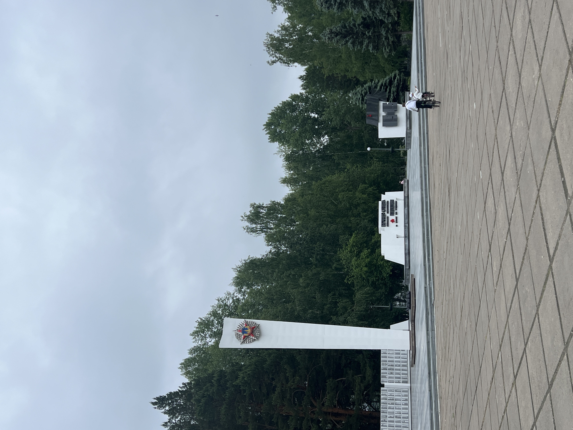 The image depicts a large war memorial complex in Chaikovsky, Russia, on an overcast day. In the foreground, a vast, light grey paved square with square concrete tiles extends towards the memorial structures.
The central feature is a tall, slender, white monument with a stylized red, blue, and gold star emblem mounted on its upper section. To the left of this monument, a long, lower white wall is visible, composed of numerous rectangular plaques or inscribed panels.
Further right, behind a lower white structure with red text (partially visible, likely a memorial inscription such as "ETERNAL GLORY TO THE HEROES WHO FELL IN BATTLES FOR THE HOMELAND"), an abstract, darker-colored monument is situated, partially obscured by trees.
Dense green trees, including both coniferous and deciduous species, form a natural backdrop behind the entire memorial complex. The sky above is uniformly light grey and cloudy.
In the mid-ground, on the paved square, two individuals are riding bicycles away from the viewer. The lead person, an adult wearing a white long-sleeved top and dark trousers, is on a dark-framed bicycle. Trailing slightly behind is a child on a smaller bicycle, wearing a white top and light trousers or shorts, with a light-colored cap.
