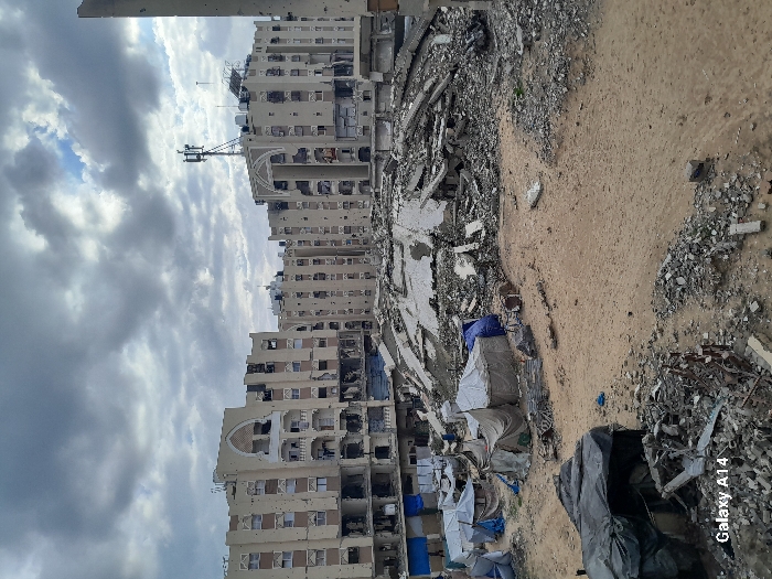 This image depicts a heavily damaged urban landscape in Khan Yunis, Palestinian Territory, under an overcast sky. In the foreground, a barren dirt ground is strewn with debris and rubble, including fragmented concrete and twisted metal. To the left, several makeshift shelters constructed from tarpaulins and fabric are clustered. The midground is dominated by a substantial mound of pulverized concrete and structural remains, indicative of a completely collapsed building. In the background, multiple multi-story residential buildings, primarily light-colored with decorative architectural elements, stand significantly damaged. These buildings exhibit numerous blast holes, shattered windows, and exposed interiors, with large sections of their facades missing or compromised. A communications tower is visible atop one of the buildings on the right. No individuals or discernible activities are present within the scene.