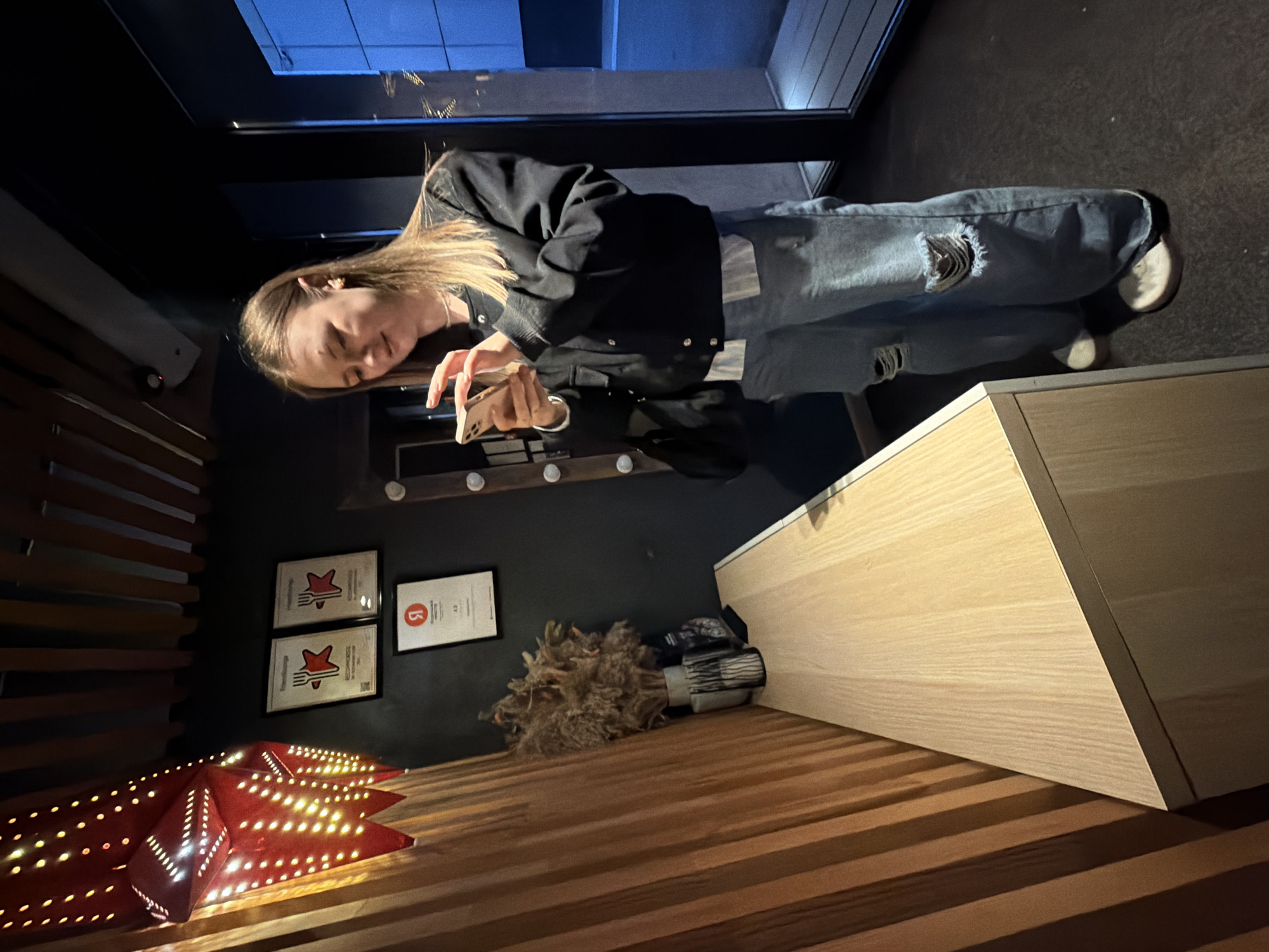 The image captures a young woman inside a dimly lit interior of a commercial establishment in Chaikovsky, Russia, possibly a restaurant or cafe. The woman, with long light brown hair, stands center-right, engrossed in her light-colored smartphone, held in both hands. She wears a black bomber jacket over a striped white shirt, wide-leg ripped light blue jeans, and light-colored flat sneakers. A black shoulder bag hangs from her left shoulder.
To her left, a large, multi-pointed red star-shaped lantern with numerous light perforations hangs, emitting a warm glow onto a vertical wooden slatted wall. Below this, a light wooden console table with a sharp angled corner extends from the left, displaying a vase with a dried floral arrangement (possibly pampas grass) and a small dark object.
On the dark gray or black wall behind the woman, three framed certificates are visible. The upper two are identical, featuring a white background with a red fork and star logo, likely indicating a "RECOMMENDED" status. Below them, a third, smaller framed certificate displays a prominent red "Я" (Yandex) logo and the rating "4.3," suggesting a review platform rating.
To the woman's right, a tall, narrow, dark-framed glass door or window reveals a dark, blue-tinted space beyond. A vertical strip of round, light-colored decorative elements, possibly part of a mirror or decorative panel, is partially visible behind her. The floor is dark and textured. The ceiling features dark wooden slats and a visible security camera or sensor.