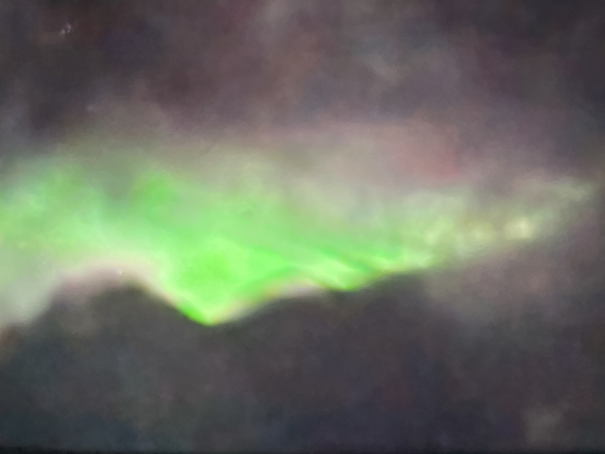The image is a close-up, abstract, and blurry shot of a bright green light against a dark, mottled background. The green light has a luminous quality and appears to be in motion, with swirling patterns and varying intensity. The background is a mix of dark grays, purples, and hints of red, creating a sense of depth and atmosphere. The overall impression is that of an ethereal or energetic phenomenon. There are no discernible objects, people, or environmental details that would suggest a specific location in a city or anywhere else.