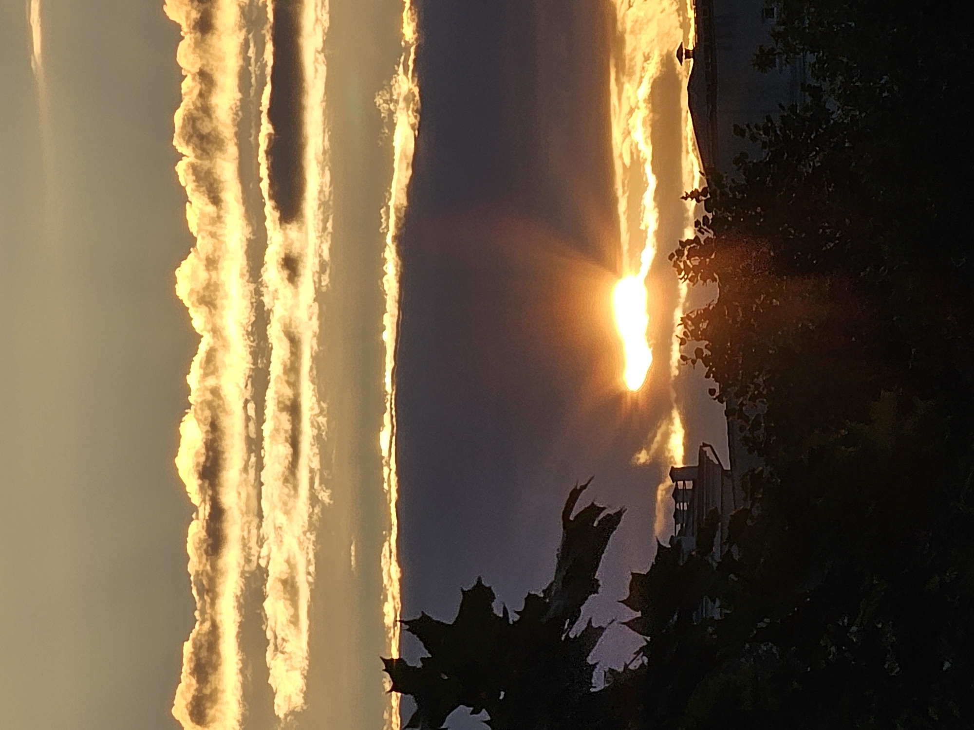 The image shows a dramatic sunset over the city of Khimki, Russia. The sun is a bright, blazing orb peeking through a band of dark clouds, casting a warm, golden light on the sky and the layered clouds above. Rays of light are fanning out from the sun, creating a halo effect. Silhouetted trees and foliage are visible in the foreground, along with the rooftops of buildings. One building in the middle ground has a distinctive turret-like structure. The overall impression is one of natural beauty and urban landscape blending together at the end of the day.