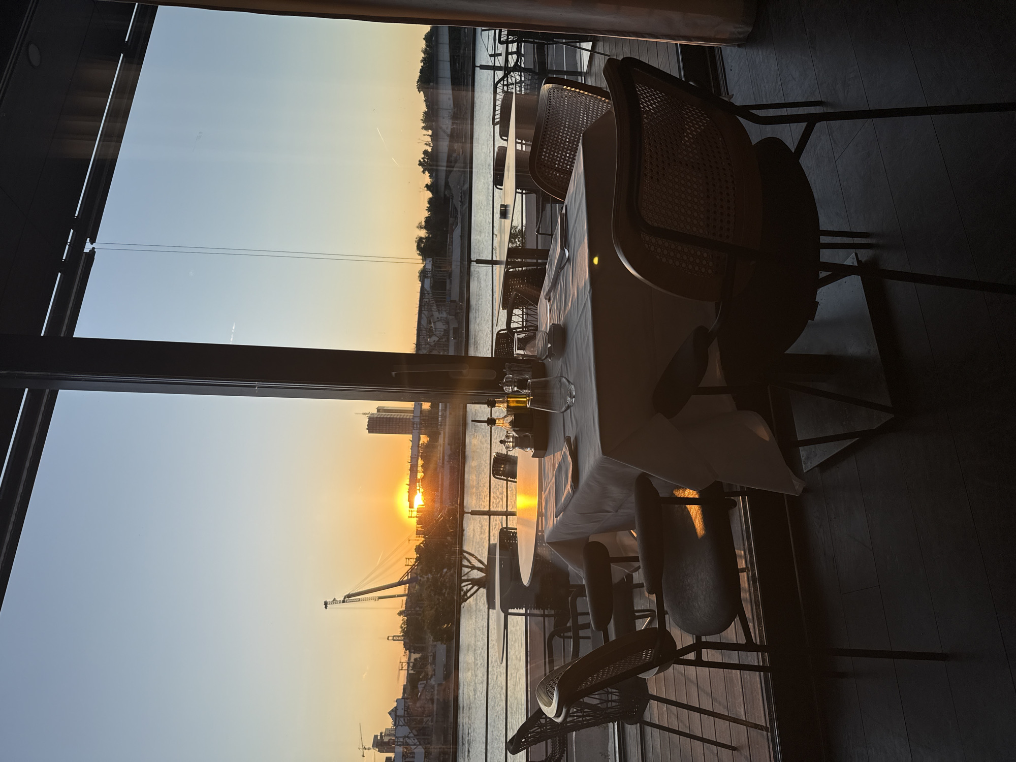 The image shows a view from inside a restaurant overlooking the Sava River at sunset in Belgrade, Serbia. In the foreground, a dining table is set with a white tablecloth, glassware, and cutlery. The chairs surrounding the table are made of dark metal frames with woven seats and backs. Beyond the table, the sunset is reflected in the water, with a construction crane visible. There is a building visible on the left bank.