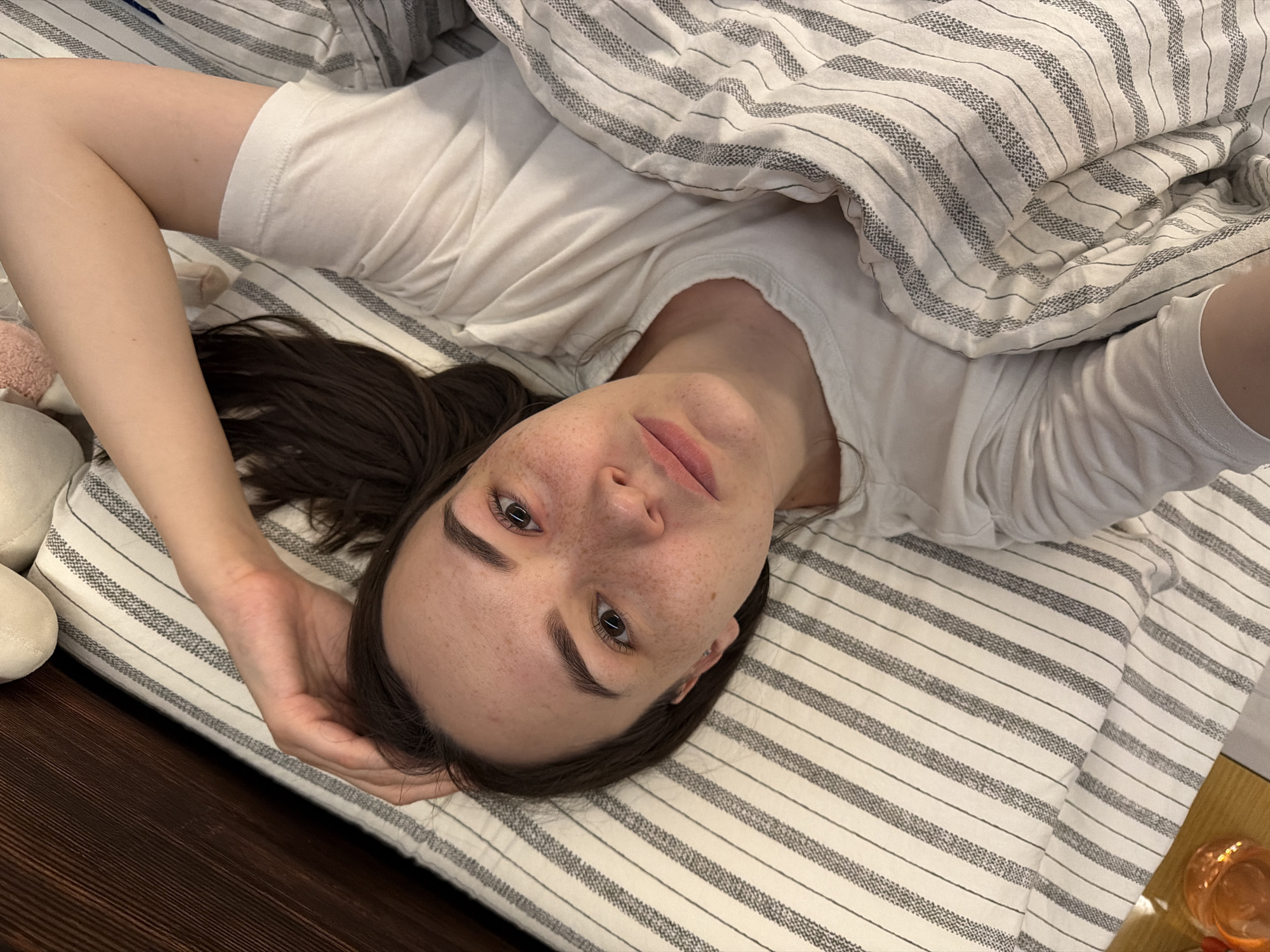 A woman is lying in bed, positioned diagonally in the frame, with her head towards the bottom left. She has dark hair, freckles, and is wearing a white t-shirt. She is partially covered by a striped duvet. Her left arm is bent and placed behind her head. The bed has a striped sheet. The bed appears to be made of wood. A dark wooden floor can be seen on the left of the image. A glass of orange liquid is located on a surface at the bottom right. The location of the image is unknown.