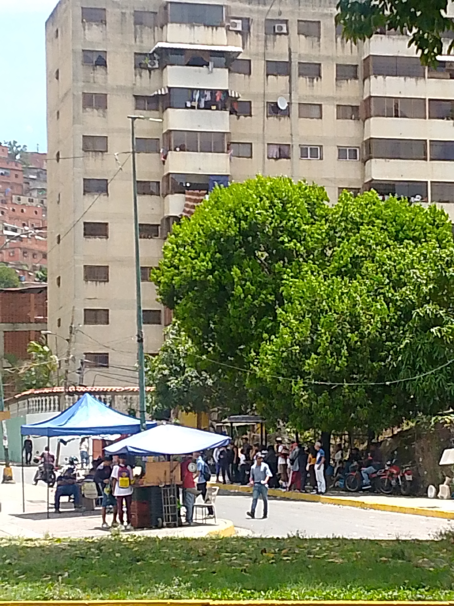 The image shows a street scene in Caracas, Venezuela, in front of a tall, weathered, multi-story apartment building. Several people are gathered near two tents with blue and white canopies. Some individuals are sitting under the blue tent, while others appear to be purchasing items from a vendor under the white tent. More people are standing along the curb, clustered near the edge of a large, green leafy tree. Several motorcycles are parked along the curb. A man in light-colored pants and a blue shirt is walking away from the crowd, across the road. The background includes a hillside covered in densely packed, red-brick buildings. A streetlight is visible near the blue tent.
