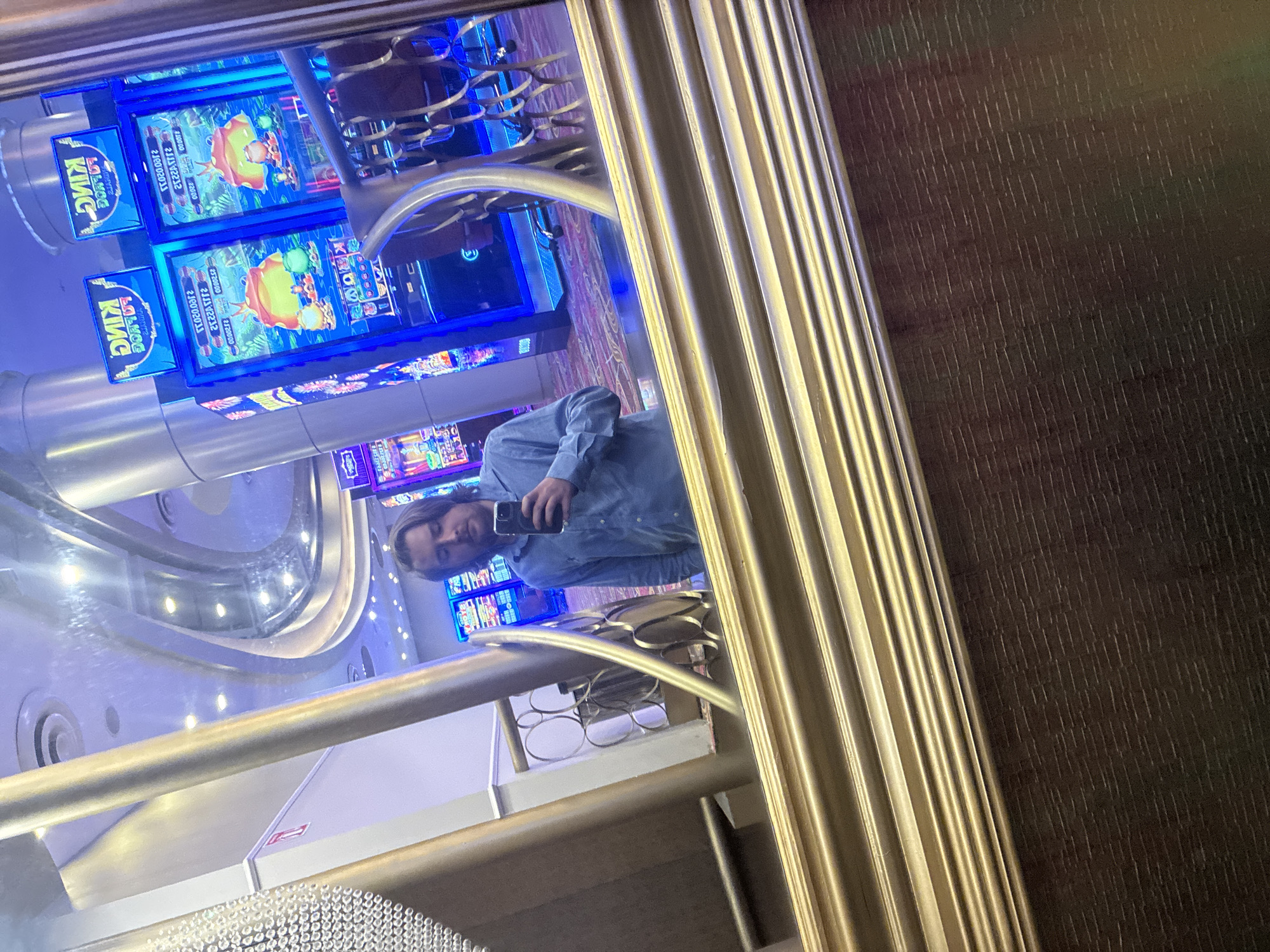 The image shows a reflection in a gold-framed mirror. A man is visible in the reflection, holding a smartphone, likely taking the picture. Behind him, the reflection shows a casino interior with slot machines. "KING" is prominently displayed on the slot machines. The architectural details reflected include curved ceilings and ornate lighting. The image was taken in the city of Huixquilucan de Degollado, Mexico.