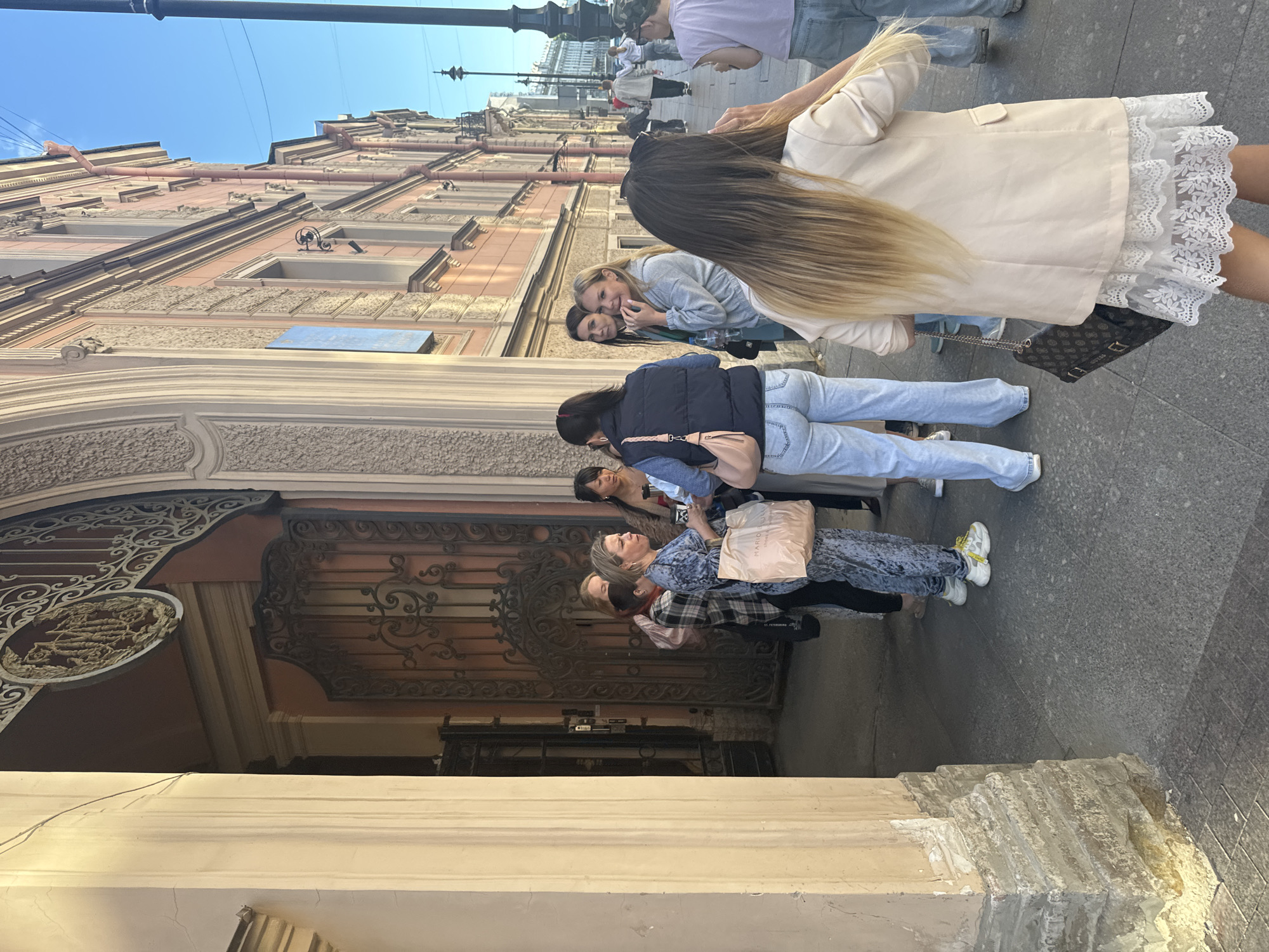A group of people are standing on a sidewalk in front of a building. The building has a decorative facade with intricate metalwork on the entrance. The people are dressed in casual clothing, and some are holding bags. The scene appears to be set in a city, possibly Saint Petersburg, Russia, given the architectural style. The lighting suggests it is daytime, with clear skies. The people seem to be engaged in conversation or waiting, with some looking at their phones. There is a sense of leisure and urban life in the image.