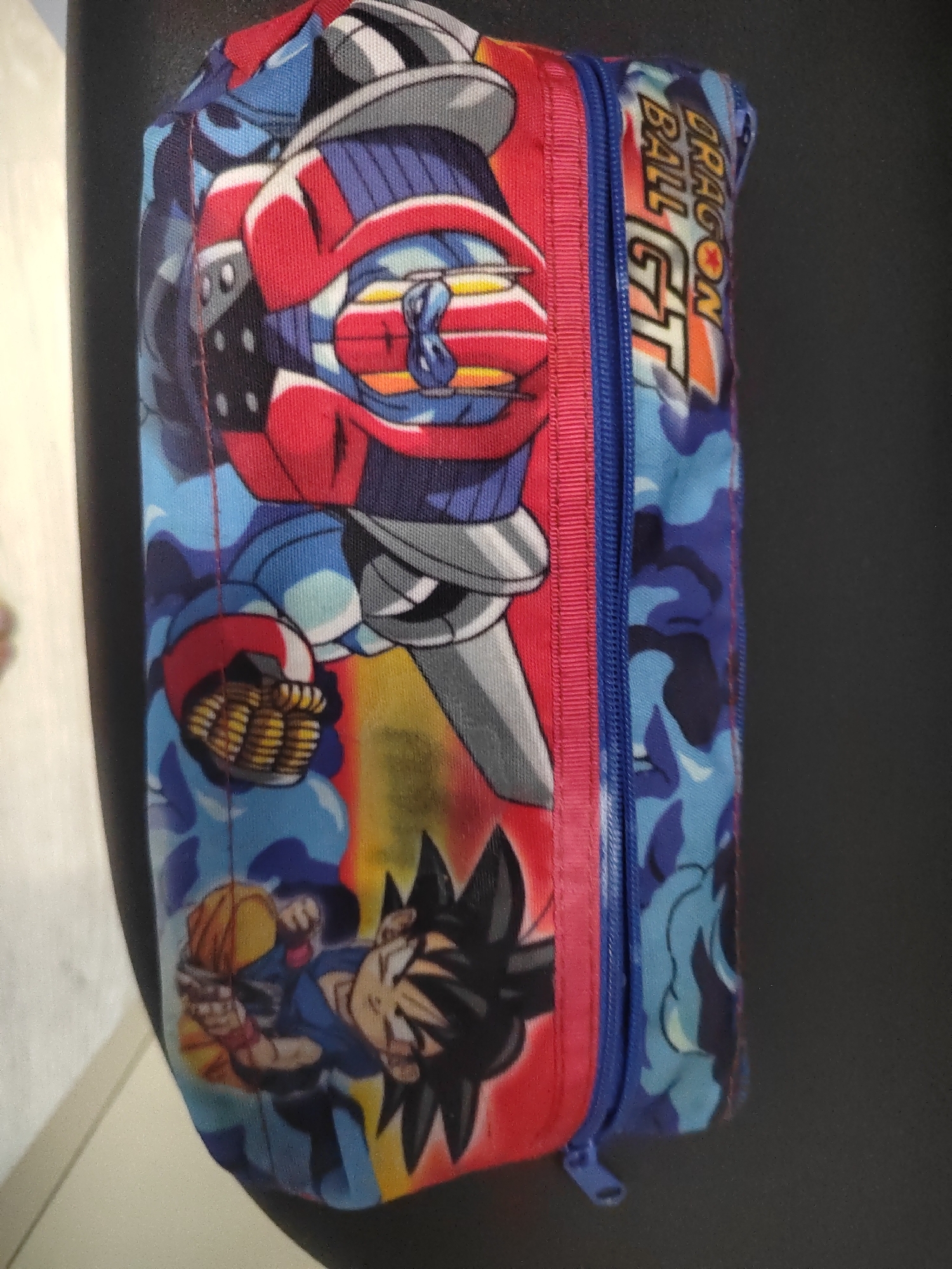 The image shows a close-up of a rectangular pencil case with a zipper. The pencil case is decorated with colorful anime characters from Dragon Ball GT. The main characters depicted are Goku and what appears to be a mecha. The fabric of the pencil case has a camouflage pattern in shades of blue and black on one side, and a vibrant mix of red, blue, yellow, and black on the other, featuring the characters. A pink ribbon runs along the length of the zipper, which is blue. The pencil case is placed on a dark, textured surface, possibly a chair or a table. The lighting suggests an indoor setting. There are no identifiable landmarks or elements that would place this image specifically in Porto Recanati, Italy.