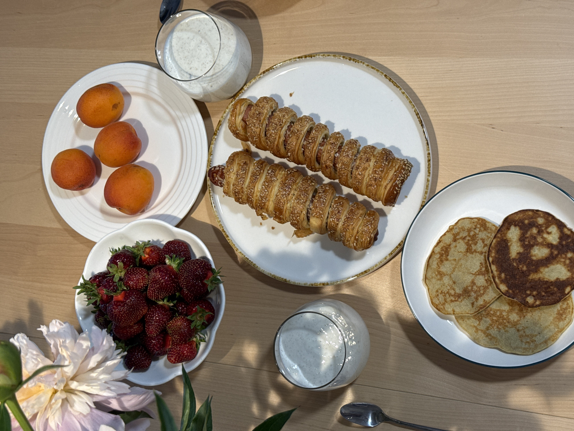 On a light-colored wooden table, a breakfast spread is laid out. In a white bowl, fresh strawberries are piled high. To the right, a white plate holds four apricots. Another white plate in the foreground displays two pastries wrapped around what appear to be sausages, sprinkled with sesame seeds. Next to this plate is a clear glass filled with a creamy, speckled beverage. Below the apricot plate, another white plate holds three round pancakes, one of which is darker. A spoon rests on the table to the left of the pancake plate. A large, light-pink peony flower with green leaves is positioned in the upper left corner of the image. The scene appears to be an indoor setting, likely a home, in the city of Izhevsk, Russia, given the context.