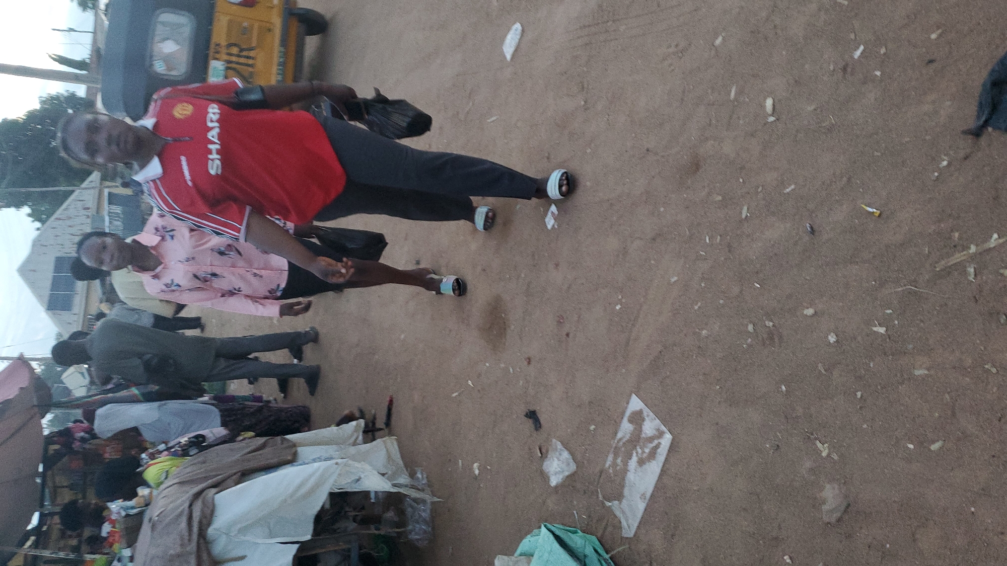 In Jalingo, Nigeria, two women walk along a dirt road lined with stalls. The woman on the left wears a pink shirt with a floral pattern, and the woman on the right wears a red Manchester United jersey with "SHARP" printed across it. Both women are carrying black plastic bags. In the background, other people are visible near market stalls and a yellow three-wheeled vehicle. The ground is dusty and scattered with debris, suggesting a typical market environment.