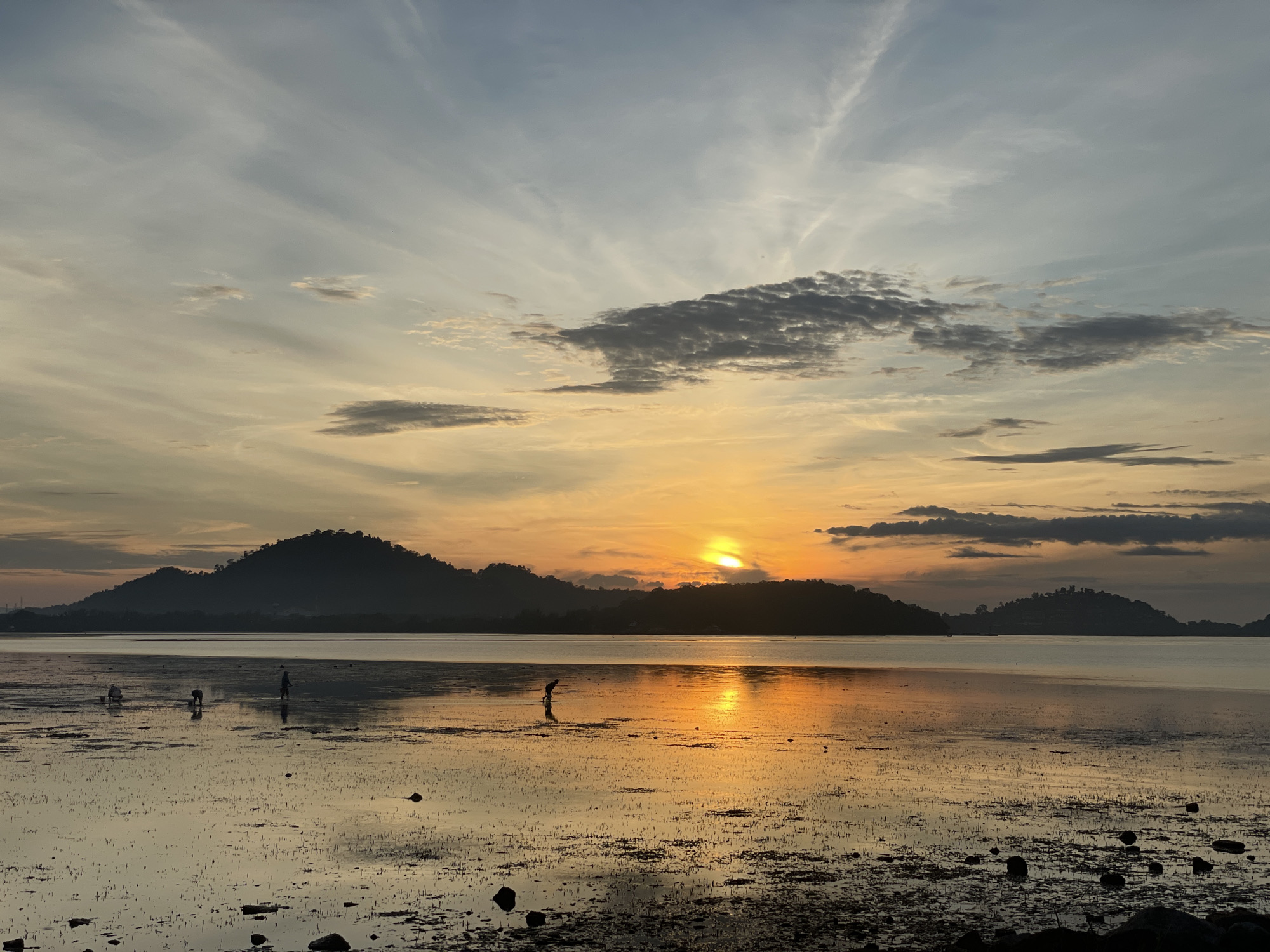 This image depicts a serene sunset over a body of water, possibly a bay or estuary, with silhouetted islands in the background. The sky is a blend of warm oranges, yellows, and soft blues, with scattered clouds. The sun, a bright orb peeking through the clouds, casts a shimmering golden reflection on the water's surface. The foreground is a shallow, muddy area with scattered rocks and vegetation, suggesting a low tide. Several figures, appearing to be people, are present in the shallow water, some bent over as if collecting shellfish or engaged in other activities. The overall scene evokes a sense of tranquility and daily life in a coastal community. While the specific location is not explicitly stated, the presence of tropical islands and the overall aesthetic are consistent with the natural beauty found in Phuket City Municipality, Thailand, particularly in areas with tidal flats and coastal landscapes during sunset.