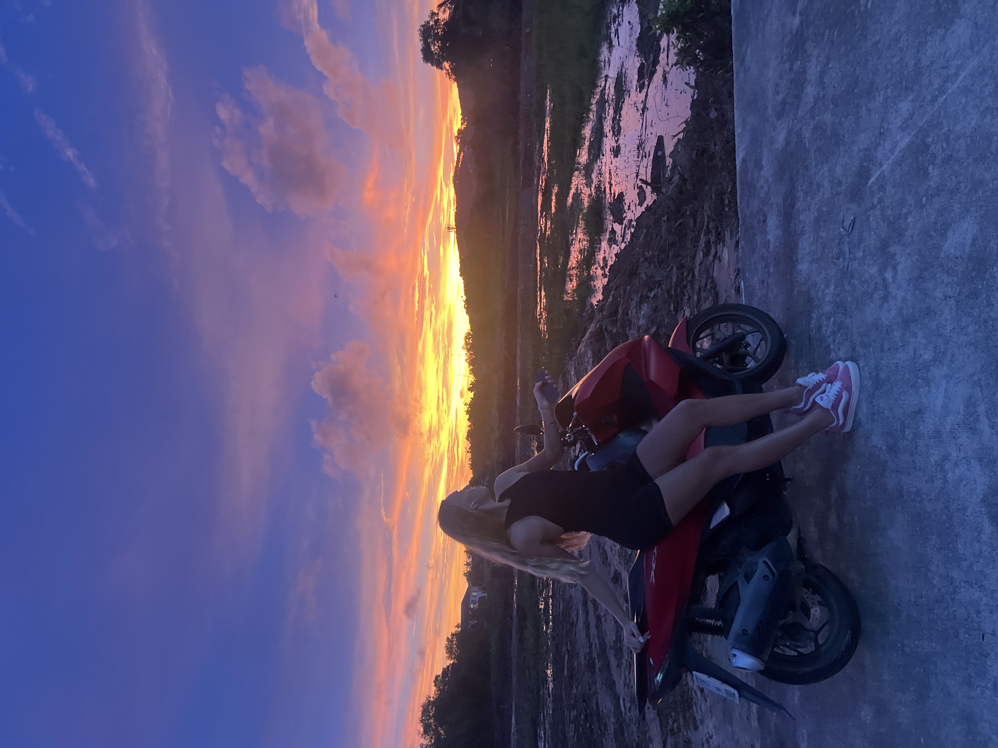 A young woman with long blonde hair is sitting on a red scooter facing right, with her left arm resting on the handlebars and her right arm extended behind her. She is wearing a black sleeveless top and black shorts, and pink Vans sneakers. The scooter is parked on a concrete surface. In the background, a vibrant sunset paints the sky with hues of purple, blue, orange, and yellow. The landscape features a body of water reflecting the colorful sky, muddy ground, and a line of trees and hills. A building and a distant cell tower are visible on the horizon. The location is Thep Krasatti, Thailand.