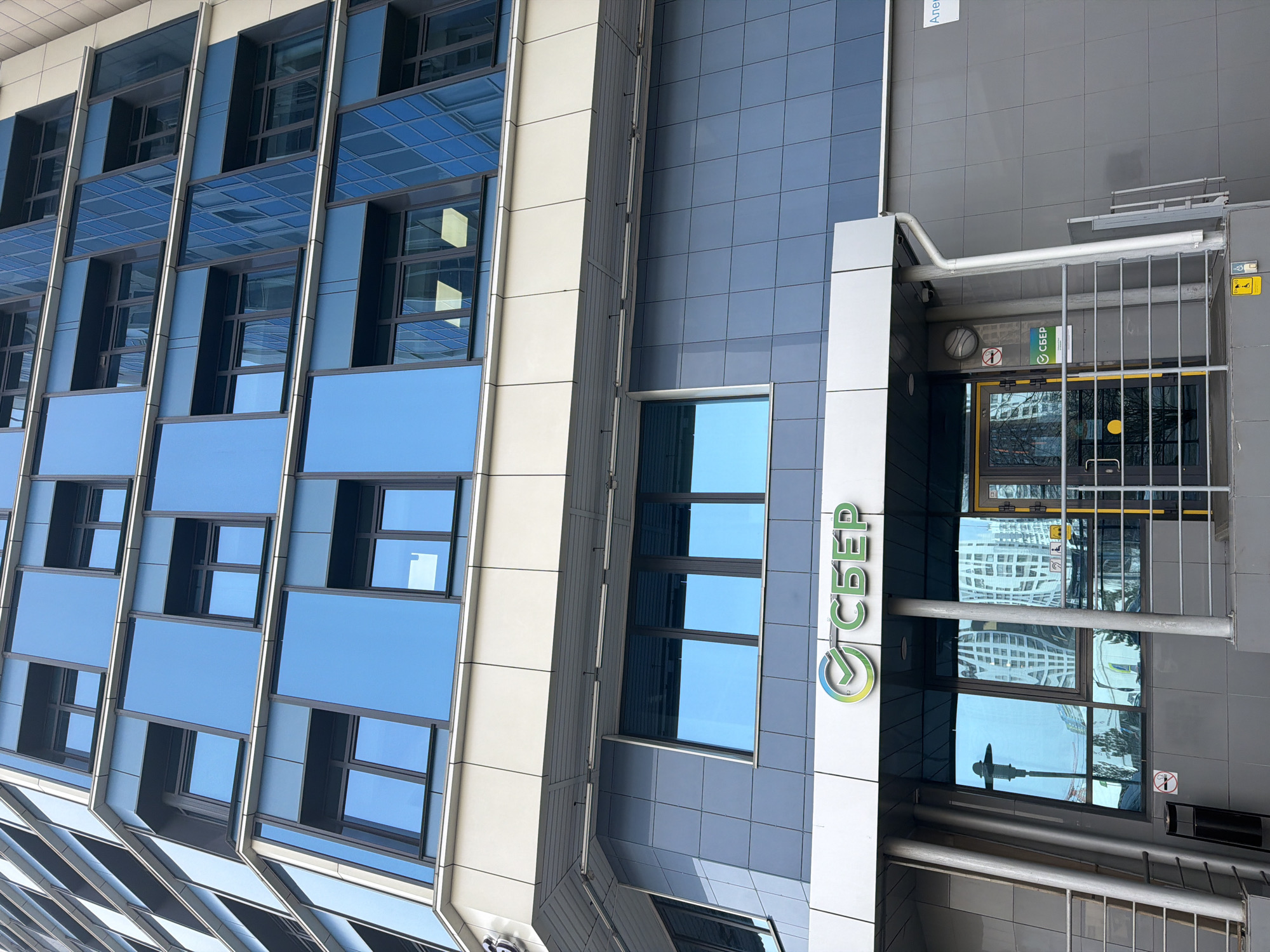 The image depicts the exterior of a multi-story building in Krasnoyarsk, Russia. The building features a modern architectural style with a facade composed of reflective blue glass panels, interspersed with darker blue and white vertical structural elements. A prominent sign with the word "СБЕР" (SBER), in Cyrillic script, is affixed to the side of the building. The entrance is visible, guarded by metal bars, with reflections of surrounding buildings and sky in the glass doors and windows. The overall environment suggests an urban, commercial district.