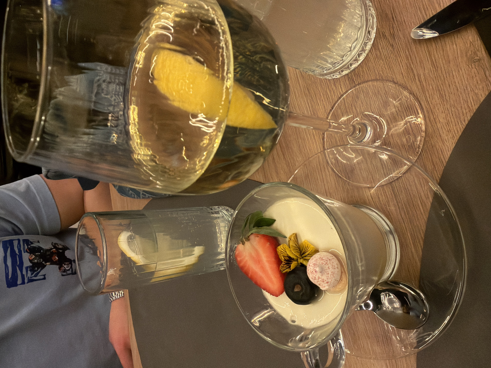 The image shows a table setting with several glasses and a dessert. Two tall glasses contain a yellow liquid, potentially lemonade or a similar beverage, with visible lemon slices inside. One is an ordinary glass while the other is a stemmed glass. Another stemmed glass contains a white dessert, possibly a mousse or pudding, topped with a strawberry slice, a yellow flower, a dark-colored sphere (possibly a berry or chocolate), and a speckled white and pink candy. A spoon is visible near the dessert glass. A table knife is also visible at the upper right corner. A person is visible wearing a blue t-shirt. The table appears to be made of wood with a dark brown accent. The location is unknown.