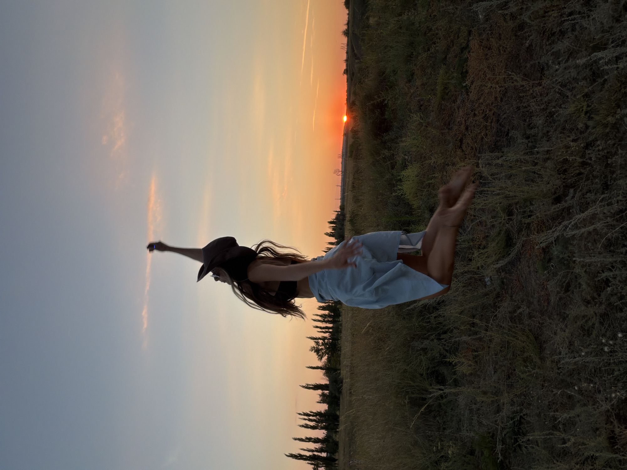 A full-body, side-profile shot captures a young woman in mid-air, jumping joyfully in a grassy field during sunset. She is wearing a dark wide-brimmed hat, a black halter top, and a light blue skirt. Her right arm is raised, and her long, dark hair trails behind her. The background features a vibrant sunset with hues of orange and pink, silhouetting a line of tall, thin trees in the distance. The field is covered in dry grass and other low-lying vegetation. The scene appears to be in an open, rural area, possibly on the outskirts of Kamyshin, Russia, given the natural landscape and absence of urban structures.