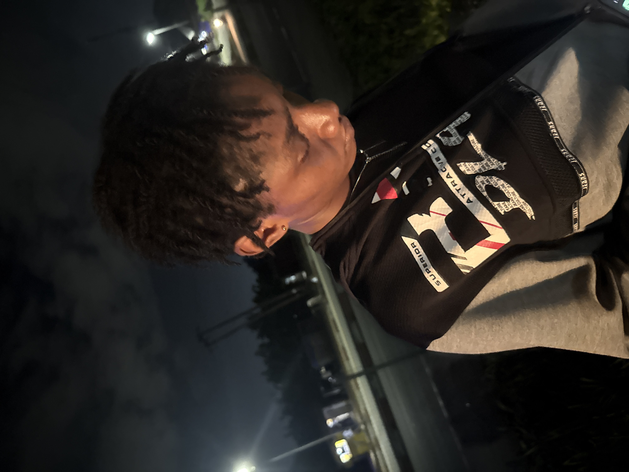 A low-angle, medium shot captures a person with dreadlocks looking down, partially illuminated by a street lamp. The individual wears a black and grey long-sleeved shirt with graphics, including the word "SUPERIOR" and other text. A silver necklace is visible around their neck. The background suggests an outdoor urban environment at night, with blurred streetlights, road markings, and trees. The scene appears to be on the side of a road in Uyo, Nigeria, indicated by the presence of a street lamp and what appears to be a roadside structure.