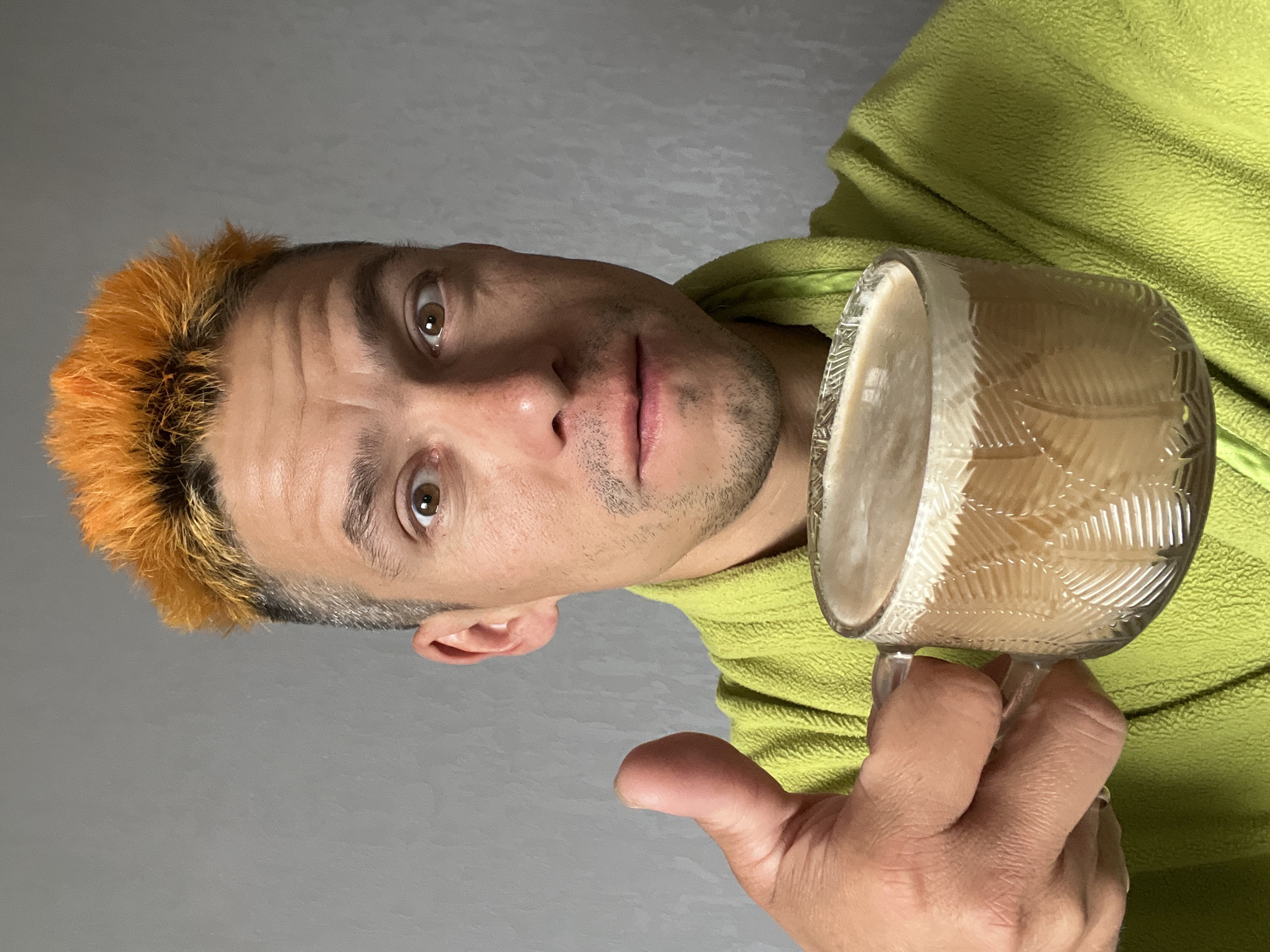 A man with short, spiky, bright orange hair and a light green hooded jacket is holding a clear glass mug filled with a light brown beverage. The mug has a textured pattern of leaves or feathers etched into its surface. The man is looking directly at the camera with a neutral expression. His eyes are light brown and he has a faint shadow of stubble on his face. The background is a plain, light gray wall, suggesting an indoor setting. The lighting is even and diffused, with no strong shadows, which could indicate it's daytime, though the specific time of day is not evident. There are no visible signs of weather conditions or specific location cues beyond the implied indoor setting. The man's thumb is visible, gripping the handle of the mug. The image is oriented sideways.