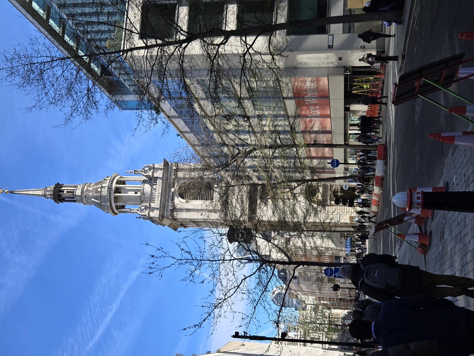 This image depicts a street scene at Cheapside in the City of London, United Kingdom, with the prominent steeple of St. Mary-le-Bow church visible towards the right-center. The scene is captured under a clear blue sky with sparse wispy clouds.
In the foreground, a paved sidewalk features several orange traffic cones, a white and red bollard, and part of a dark-suited pedestrian in the lower left. To their right, another pedestrian wearing a dark jacket and a grey backpack walks away from the camera. The street, an asphalt road, runs through the center, showing traffic lights and signs further down.
To the right, a modern multi-story building with a glass and light-colored facade is visible. Its lower section displays a red banner with white text that partially reads "More Whistles." Construction barriers and ladders are present near its ground-level entrance. Bare deciduous trees are interspersed along the street and in front of buildings.
Numerous pedestrians are observed on both sidewalks and crossing the street, indicating a busy urban environment. Beyond the immediate foreground, a line of diverse city buildings, both historic and contemporary, recede into the distance along the street. Shadows suggest a sunny day.
