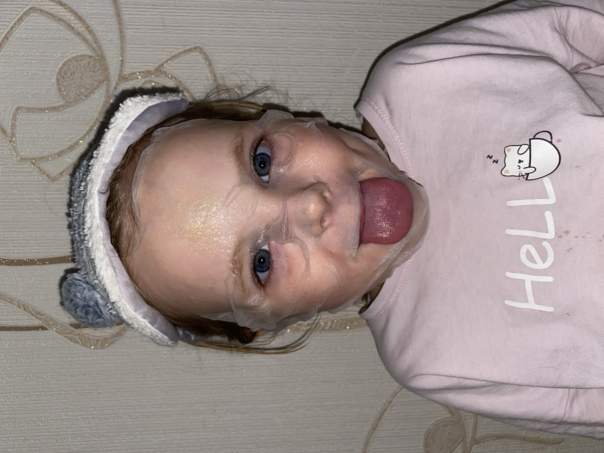 An overhead shot, rotated 90 degrees counter-clockwise, shows a young child lying down indoors. The child is wearing a transparent sheet face mask that covers their entire face, with cutouts for the eyes, nose, and mouth. Their bright blue eyes are wide open, and their tongue is playfully sticking out.
The child has light brown hair and is wearing a fuzzy gray and white headband with ear-like embellishments on top. They are dressed in a light pink long-sleeved shirt or sweatshirt. On the shirt, a graphic of a white cartoon cat sleeping in a white mug is visible, accompanied by "Zz" symbols indicating sleep. Below the graphic, the word "Hello" is partially visible in white lettering. The shirt also shows some light stains or marks.
The background consists of a light-colored, possibly off-white or cream, textured wallpaper featuring a shimmering, intricate floral or abstract swirl pattern in a light brown or gold hue. The lighting appears to be artificial and warm. The overall scene suggests a playful and relaxed moment, possibly during a skincare routine or dress-up play.