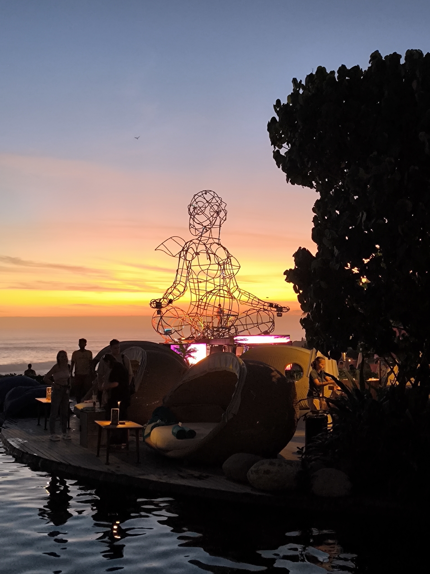 The image depicts a twilight scene at a beachfront establishment in Kediri, Indonesia. An elevated platform appears to extend into a pool of water, the surface rippling. On the platform, several people are present, standing near tables. One woman is partially visible in the center foreground, along with two men standing behind her. Other individuals can be seen in the background near lounge chairs. Dominating the scene is a large, abstract wire sculpture of a meditating figure placed atop a structure of lounge chairs. The sculpture is silhouetted against a vibrant sunset displaying orange and yellow hues. To the right, a dark tree partially frames the composition. In the distant sky, a small bird appears in flight. The scene appears to be a social gathering, likely a bar or restaurant, at dusk.