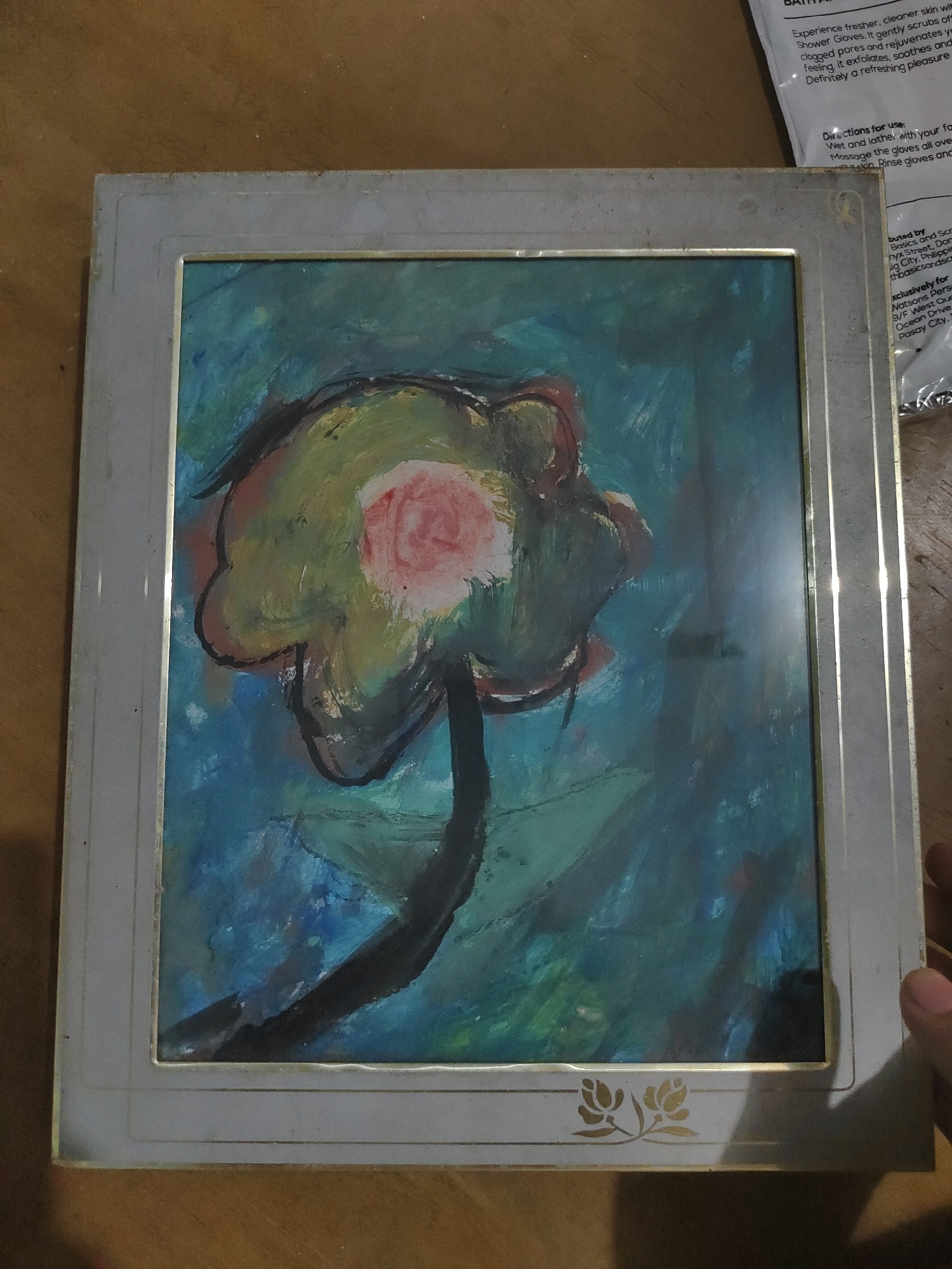 A framed painting depicting a stylized flower with a pink center, surrounded by abstract green, yellow, and brown petals, outlined in black, rests on a dark stem against a blue and green background. The painting is encased in a rectangular, light grey frame with a gold inner border and a decorative golden floral emblem at the bottom. The frame shows surface wear and dust accumulation. Reflections are visible on the glass covering the artwork. The framed object is situated on a wooden surface with a visible grain. In the upper right background, a white plastic package is partially visible, displaying text related to "Shower Gloves" and instructions for use. This packaging identifies "Basics and Scrubs" as a distributor operating from Davao City, Philippines. A human hand, specifically a thumb and forefinger, is visible at the bottom right corner, holding or resting near the frame.