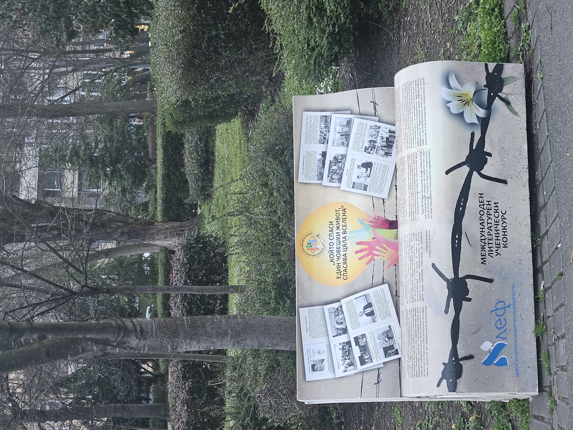 A sideways eye-level shot depicts an outdoor book display, likely in Burgas, Bulgaria, based on the Cyrillic text. The book lies open, one page depicting a white flower and barbed wire, with the text “МЕЖДУНАРОДЕН ЛИТЕРАТУРЕН УЧЕНИЧЕСКИ КОНКУРС” (International Literary Student Contest). Another page has the text “„КОЙТО СПАСИ ЕДИН ЧОВЕШКИ ЖИВОТ, СПАСЯВА ЦЯЛА ВСЕЛЕНА" ("Whoever saves a human life, saves the whole universe"). It also has a yellow circle with a logo and multi-colored hands. Several smaller printed sheets, featuring black and white photographs and text, are displayed on top of the book. The display is set on a paved surface surrounded by grass and vegetation. In the background, trees and shrubs are visible, providing depth to the outdoor setting.