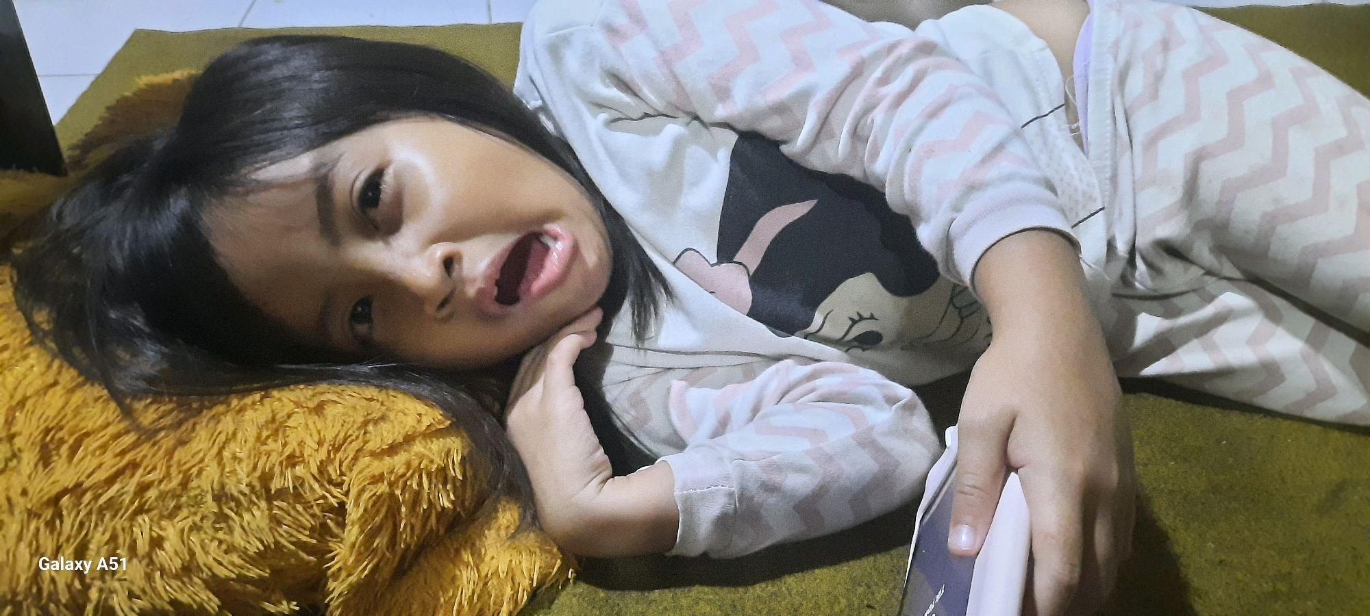 A young Indonesian girl is lying on her left side on a fluffy, mustard-yellow pillow. She is looking towards the camera with her mouth open, her right hand resting on her chin and neck. She is wearing a light-colored pajama set with a pink chevron pattern and a cartoon character graphic on the shirt. The background appears to be a floor covered with a dark green rug. The image is a close-up shot, focusing on the girl's face and upper body. The bottom left corner of the image shows the text "Galaxy A51". The scene is indoors, with an overhead light source illuminating the girl.