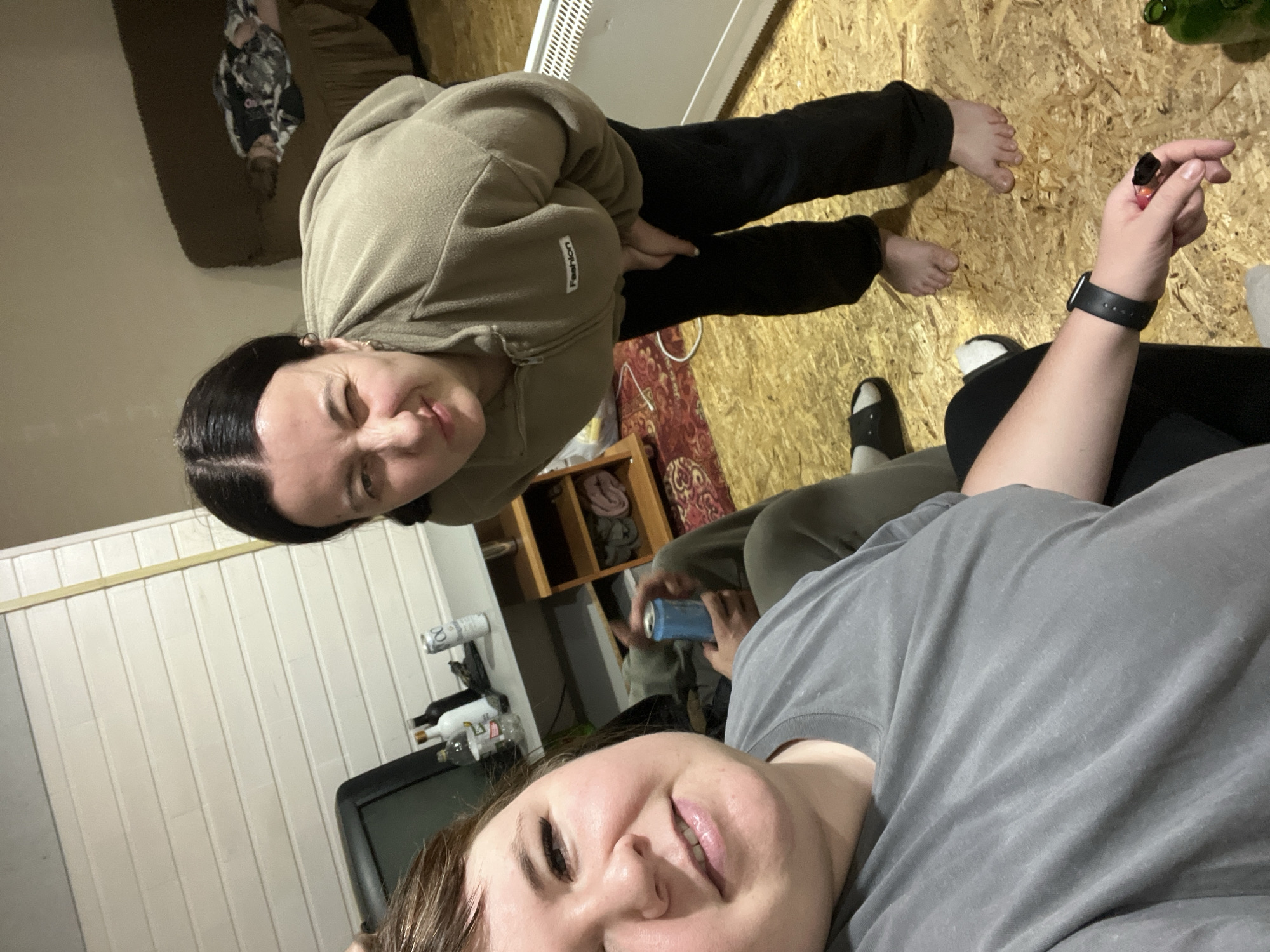 Two people are in an indoor location. One person is facing the camera, wearing a gray t-shirt. The other person is wearing a beige hoodie and black pants, facing away from the camera and looking towards the person in the gray t-shirt. A can is being held by a person. The location appears to be a room with wood flooring, a heater, a TV, shelves, wine bottles, and a couch. It is not possible to confirm the city of Chaikovsky, Russia from the image.