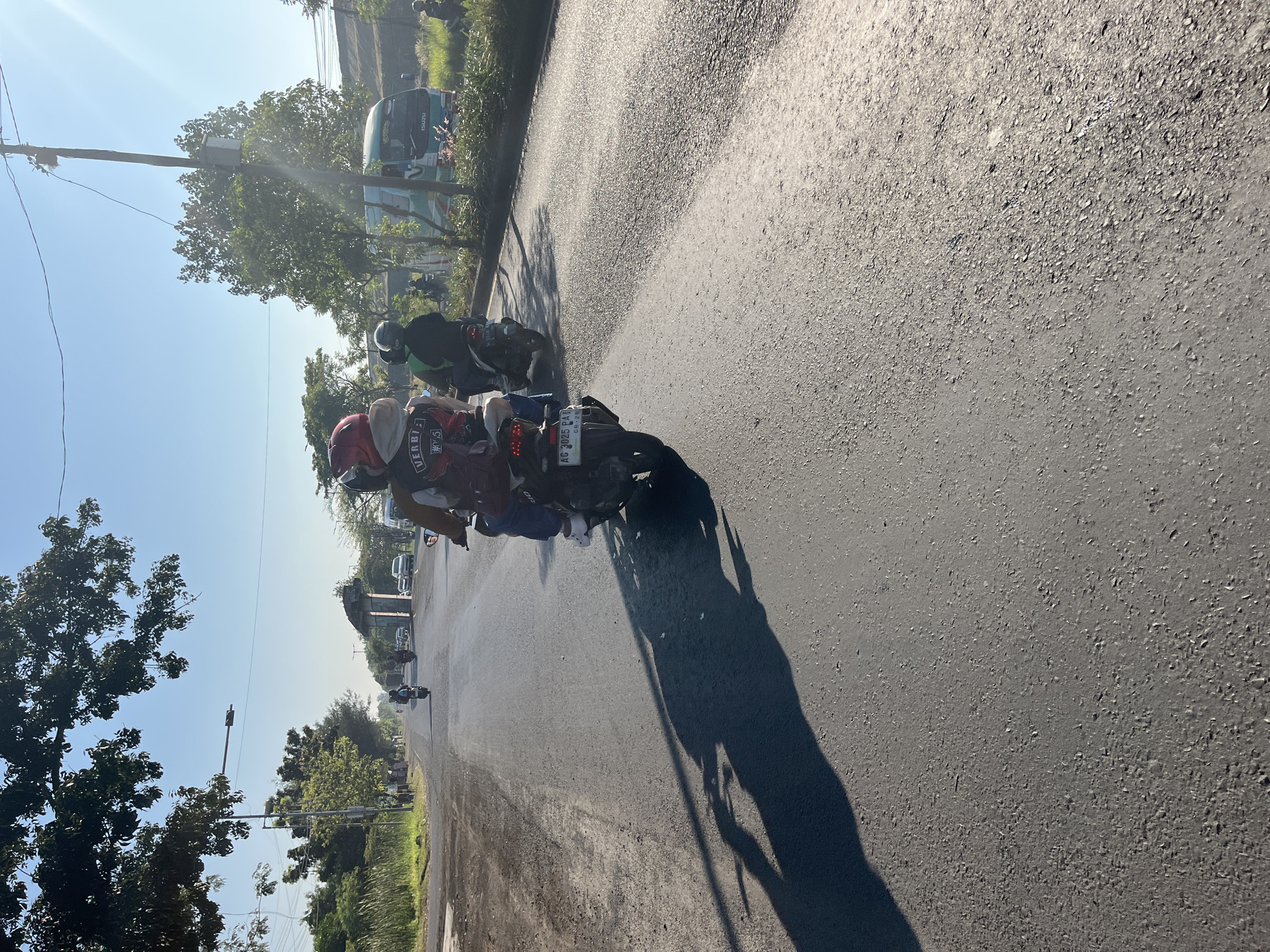 In Siring, Indonesia, on a sunny day, two people are riding a motorcycle on a paved road. The driver, wearing a red and black helmet, is partially obscured, while the passenger, in a maroon jacket, is clearly visible. The motorcycle has a license plate that reads "AG 3025 PAT". In the background, several other motorcycles and a bus are visible on the road. The scene is set against a backdrop of trees and a clear blue sky, with long shadows cast on the road, indicating it is late afternoon. The overall impression is that of a typical daily commute or journey in a developing country.