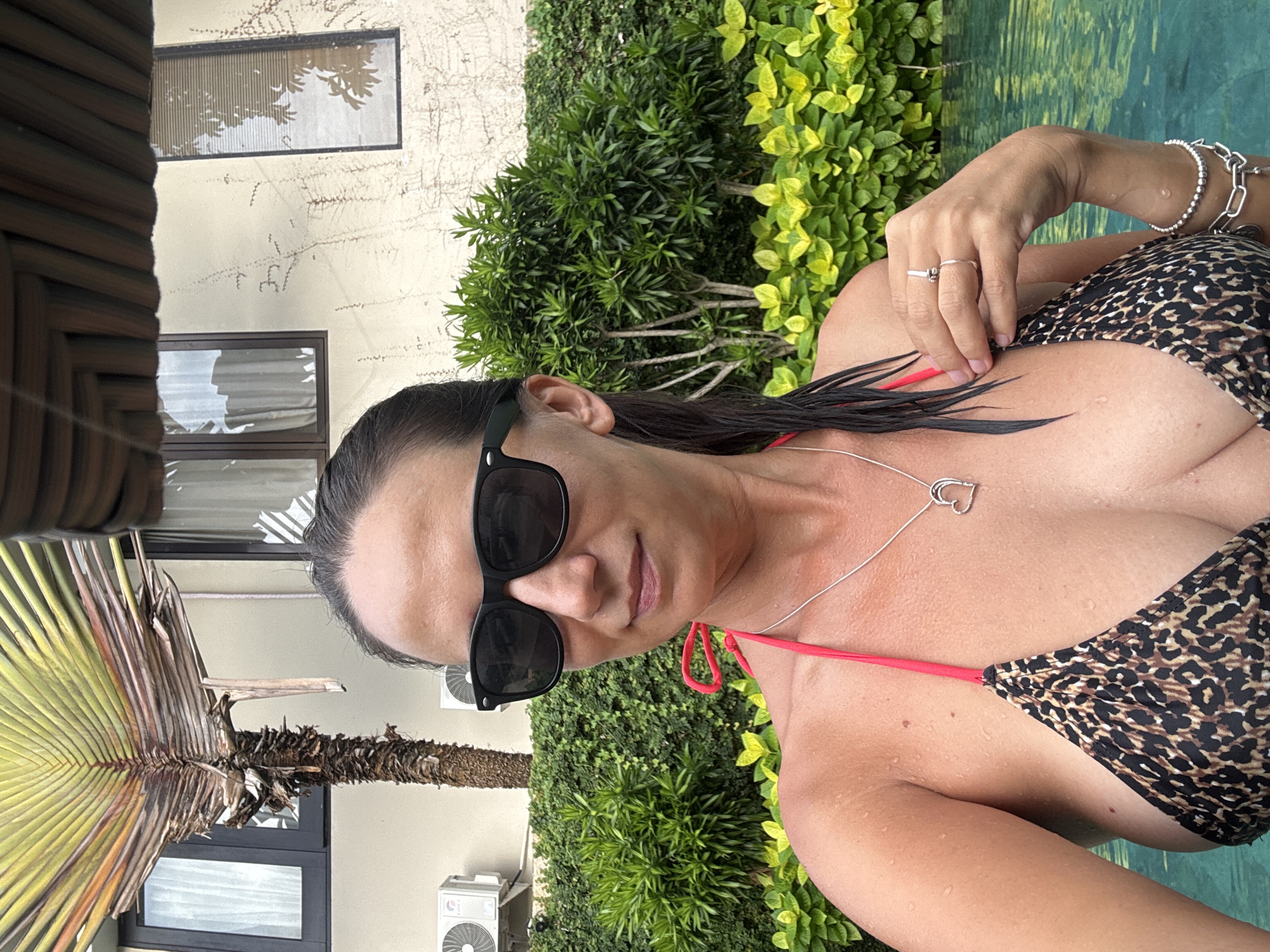 A woman is in a swimming pool with greenery behind her. She is wearing sunglasses and a leopard print bikini with red straps and a heart pendant necklace. She also has several bracelets and a ring on her finger. In the background are buildings and a palm tree. The location is Kuta Selatan, Indonesia.