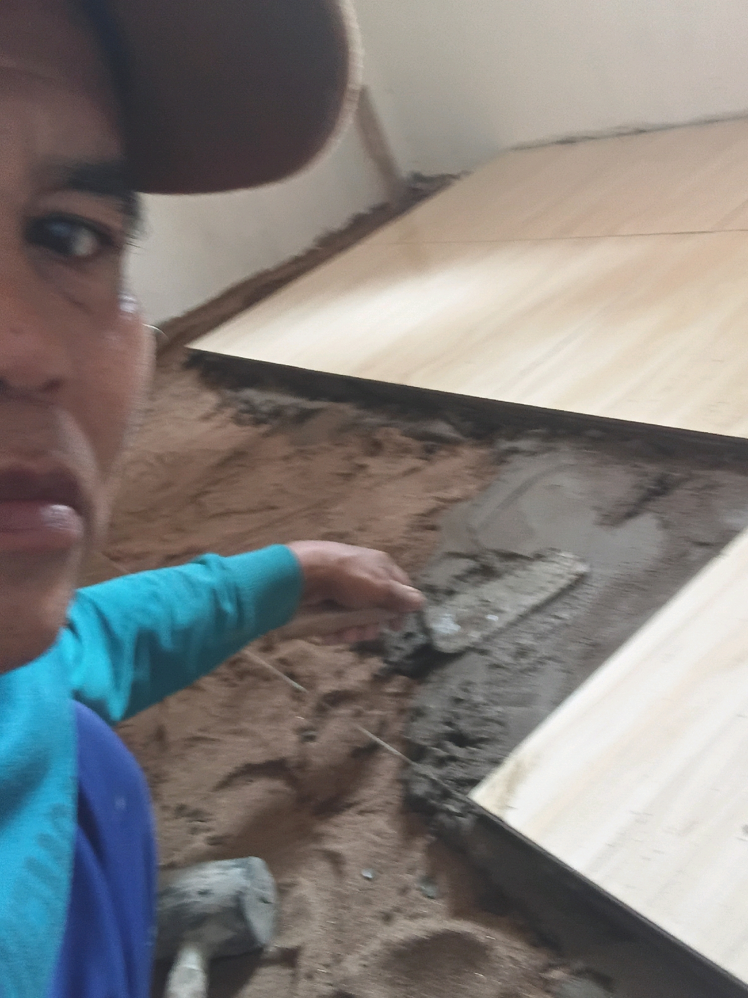 The image shows a close-up of a person's face and arm, who is engaged in construction work. The person is wearing a brown cap and a teal shirt. Their right hand is visible, holding a trowel and applying cement to a gap between two wooden surfaces. The background appears to be an indoor construction site with sand and wooden planks. The lighting suggests it is daytime. The location is identified as Gilirang, Indonesia.
