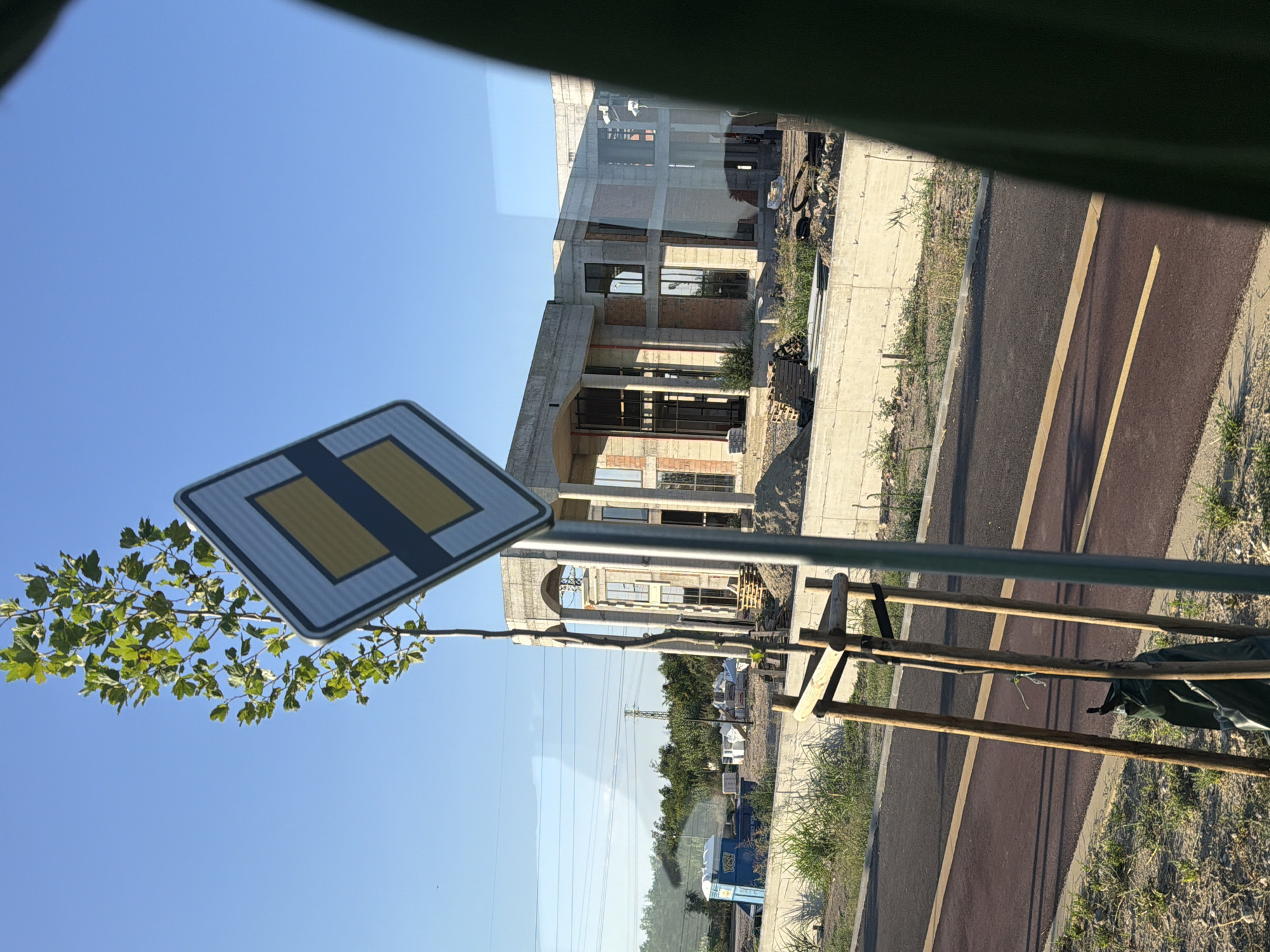 The image shows a construction site on a clear day. In the foreground, a diamond-shaped road sign with a yellow and blue pattern is visible, along with a young tree supported by wooden stakes. A paved path with yellow markings runs parallel to the construction area. In the background, a large, unfinished building with a concrete structure and brickwork is under construction. Several portable toilets are also present on the site, along with construction materials like pallets and piles of sand. The scene is likely part of a larger urban development project in Plovdiv, Bulgaria. The image captures a typical view of ongoing construction in a semi-urban or developing area.