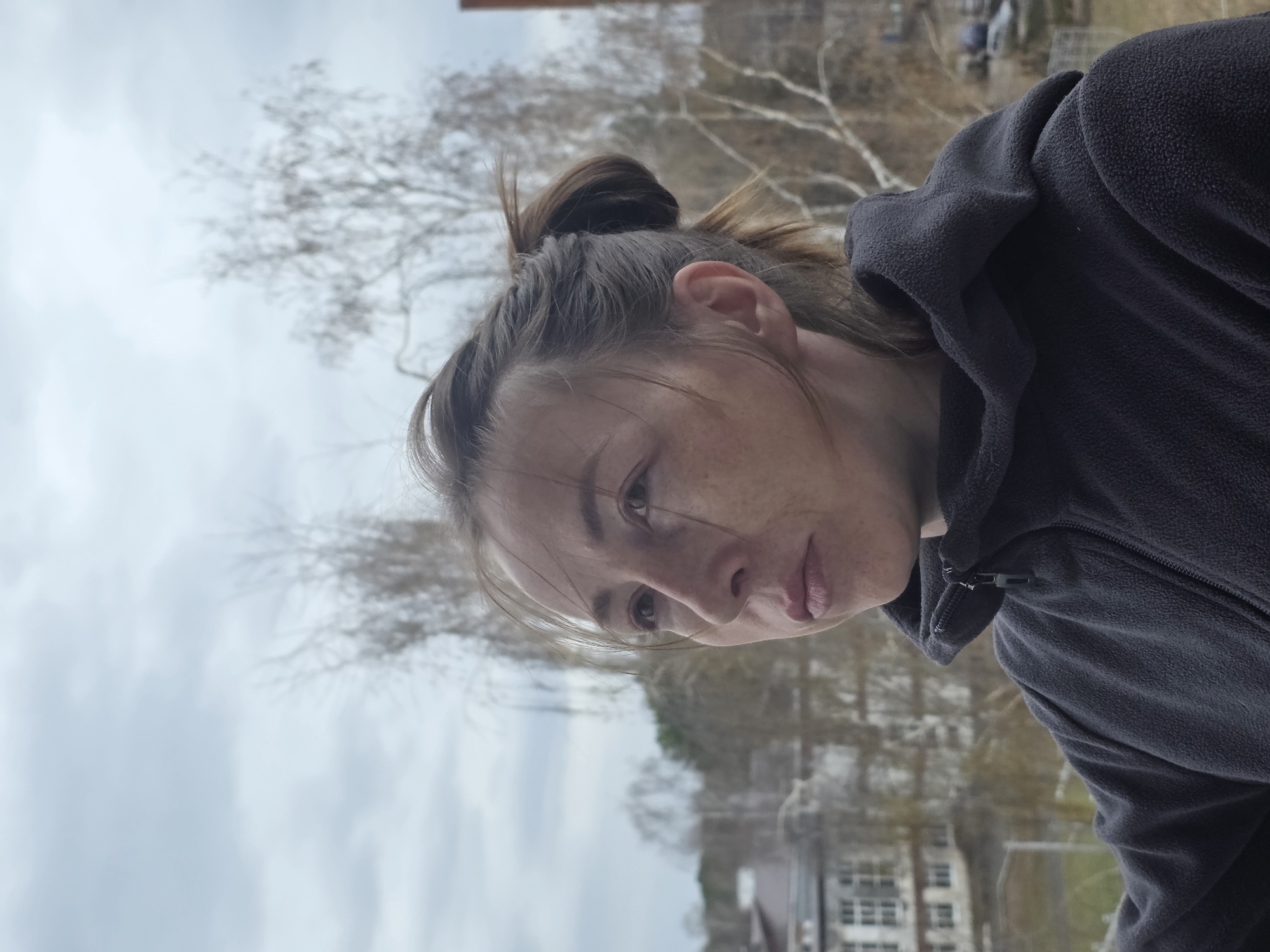 A rotated, close-up portrait captures an individual outdoors, likely in Chaikovsky, Russia. The subject is a person with brown hair tied in a messy bun, visible on the top of their head. They have scattered freckles on their face and are wearing a dark grey or black fleece jacket with a partial zipper at the collar. The person's face is turned slightly to the side, with their gaze directed downwards and to the right of the frame, and their lips slightly parted in a neutral expression.
The background is softly blurred, indicating a shallow depth of field. It depicts an overcast, light grey sky dominating the upper left portion of the scene. Bare deciduous trees, characteristic of late winter or early spring, are scattered throughout the mid-ground. Faint outlines of buildings are visible in the distant background on the right. The lighting is soft and diffused, consistent with a cloudy daytime setting. No specific activity beyond posing or observing is evident. There is no visible text in the image.