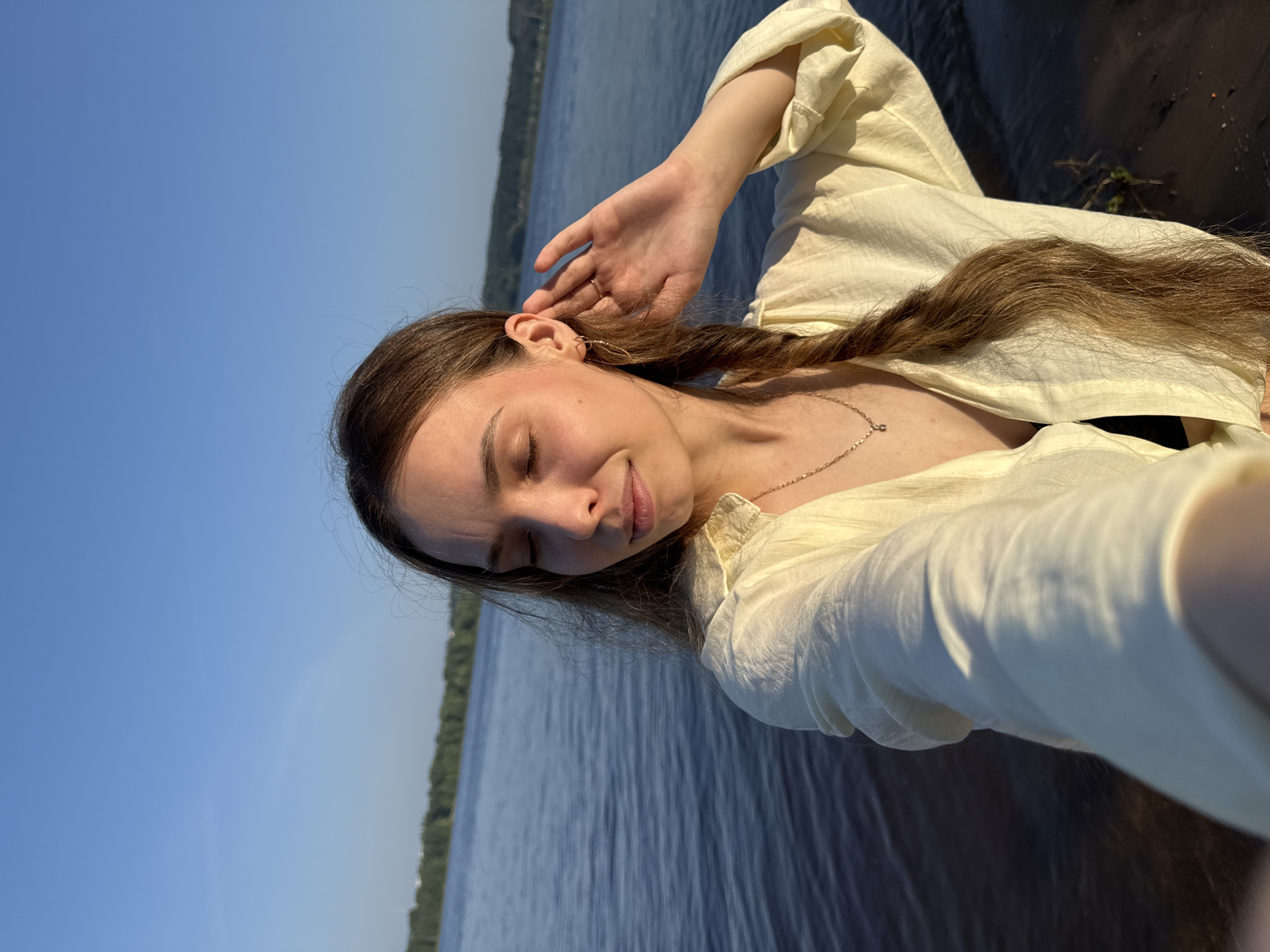 A young woman is captured in a close-up, selfie-style photograph taken outdoors during the golden hour. She stands at the edge of a body of water, possibly a lake or river, with a tree line visible in the distance across the water. The sky above is clear and a bright blue. The woman has long, light brown hair styled in a braid that hangs over her shoulder. Her eyes are closed, and she is touching her ear with her right hand, her expression suggesting a moment of reflection or relaxation. She is wearing a light yellow, unbuttoned shirt over a dark top, and a delicate necklace with a small pendant. The sun casts a warm glow on her face and skin. The water behind her is calm, with gentle ripples visible. The foreground shows a dark, sandy or muddy shore. The location is identified as Noviy, Russia.