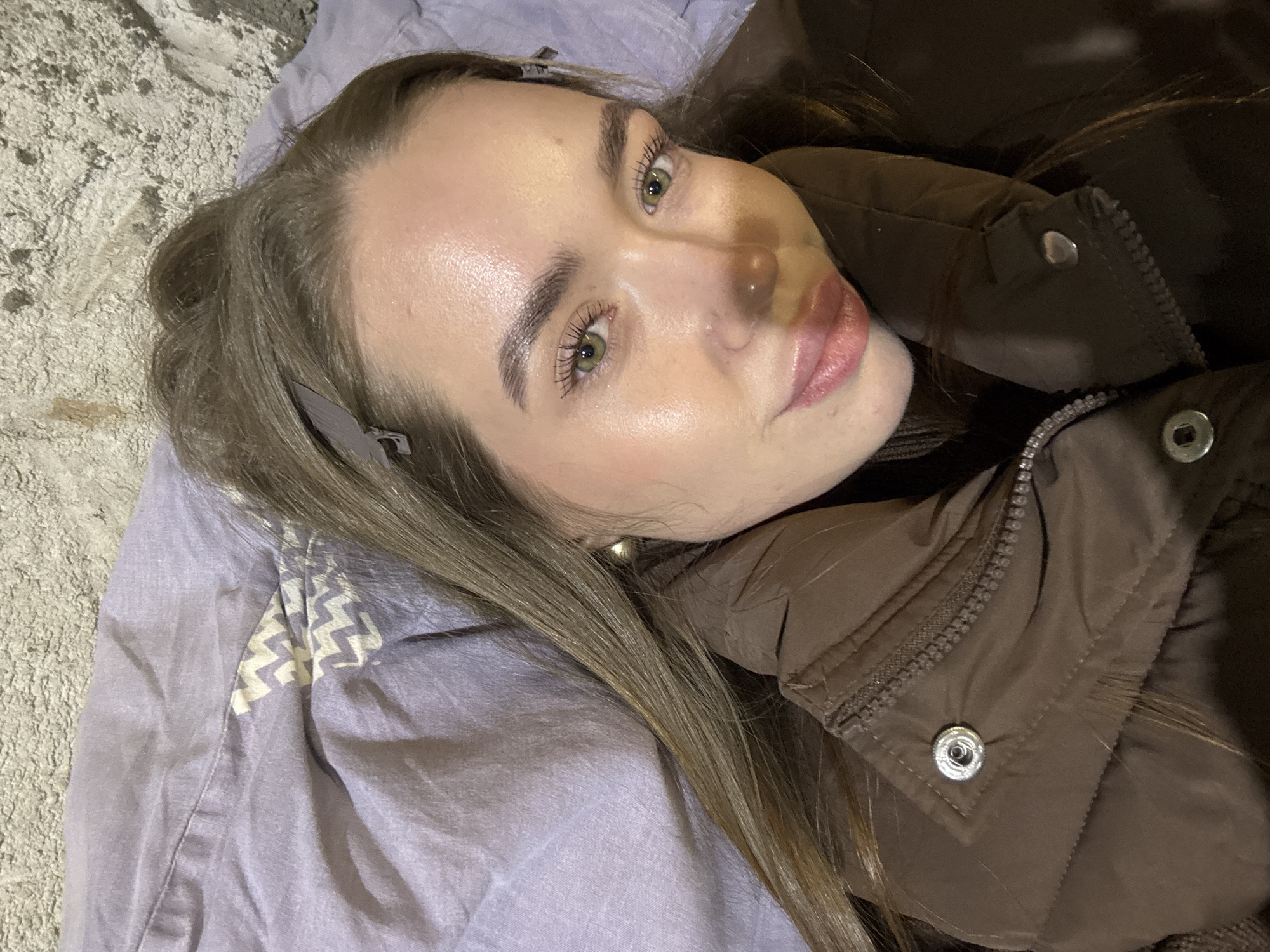 A young woman lies on her back, likely on a lilac-colored bed sheet. She has light brown hair with a hair clip and is wearing a brown puffy jacket. Her gaze is directed towards the viewer. The background is obscured but appears to include a textured, light-colored surface, possibly a wall. There is no information to support the specific location of Chaikovsky, Russia, in the image.