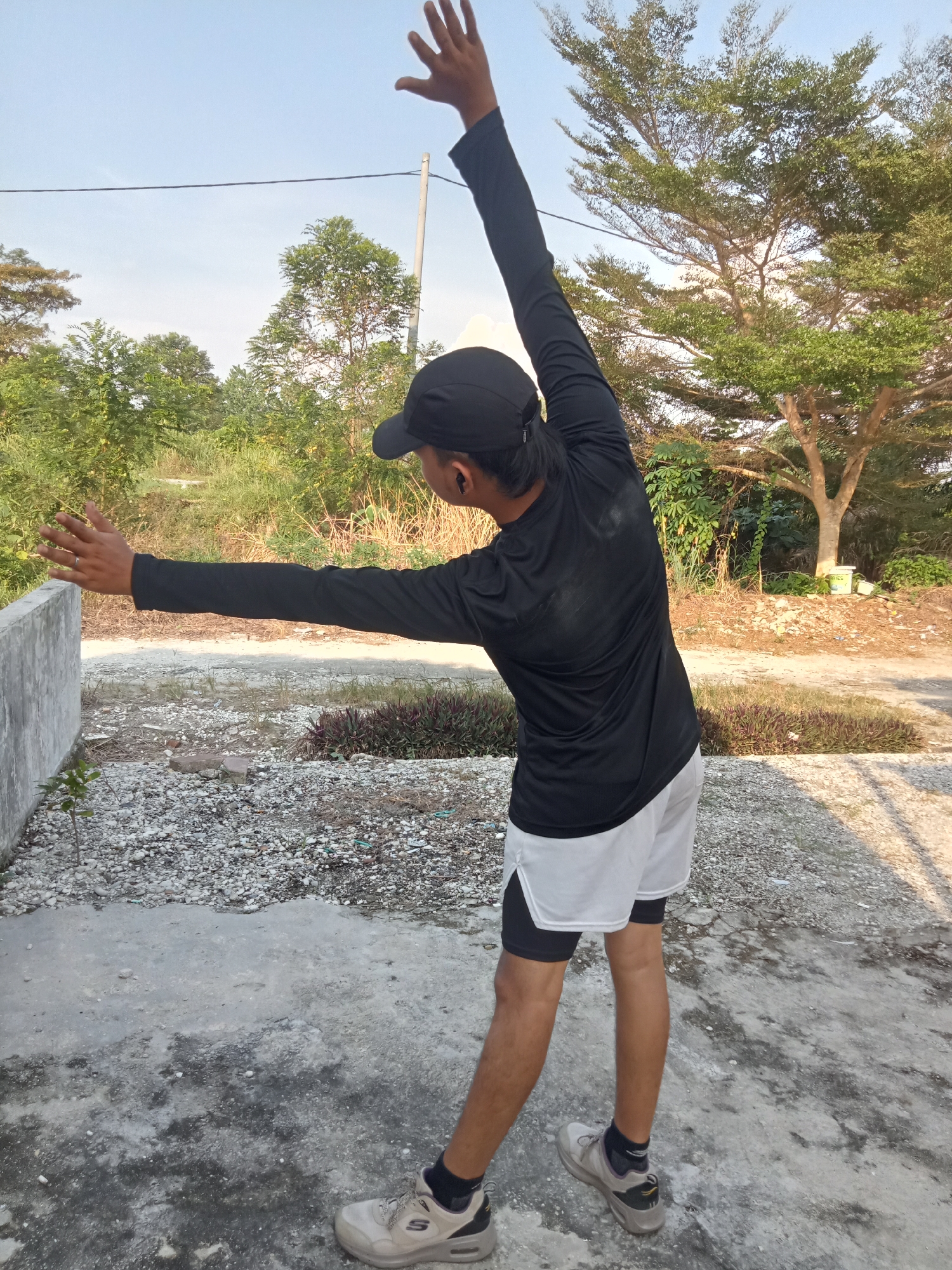 A person is performing a side stretch outdoors in Kualu, Indonesia. The individual, viewed from behind, wears a black long-sleeved shirt, white athletic shorts with black compression shorts underneath, a black baseball cap, and athletic shoes. Their left arm is extended upwards and bent at the elbow, while their right arm is extended outwards, parallel to the ground. The ground is a mixture of concrete and gravel. In the background, there are various trees and foliage, consistent with a natural or semi-rural environment within Kualu. A concrete wall is visible to the left of the frame. The sky is clear and blue, indicating daylight.