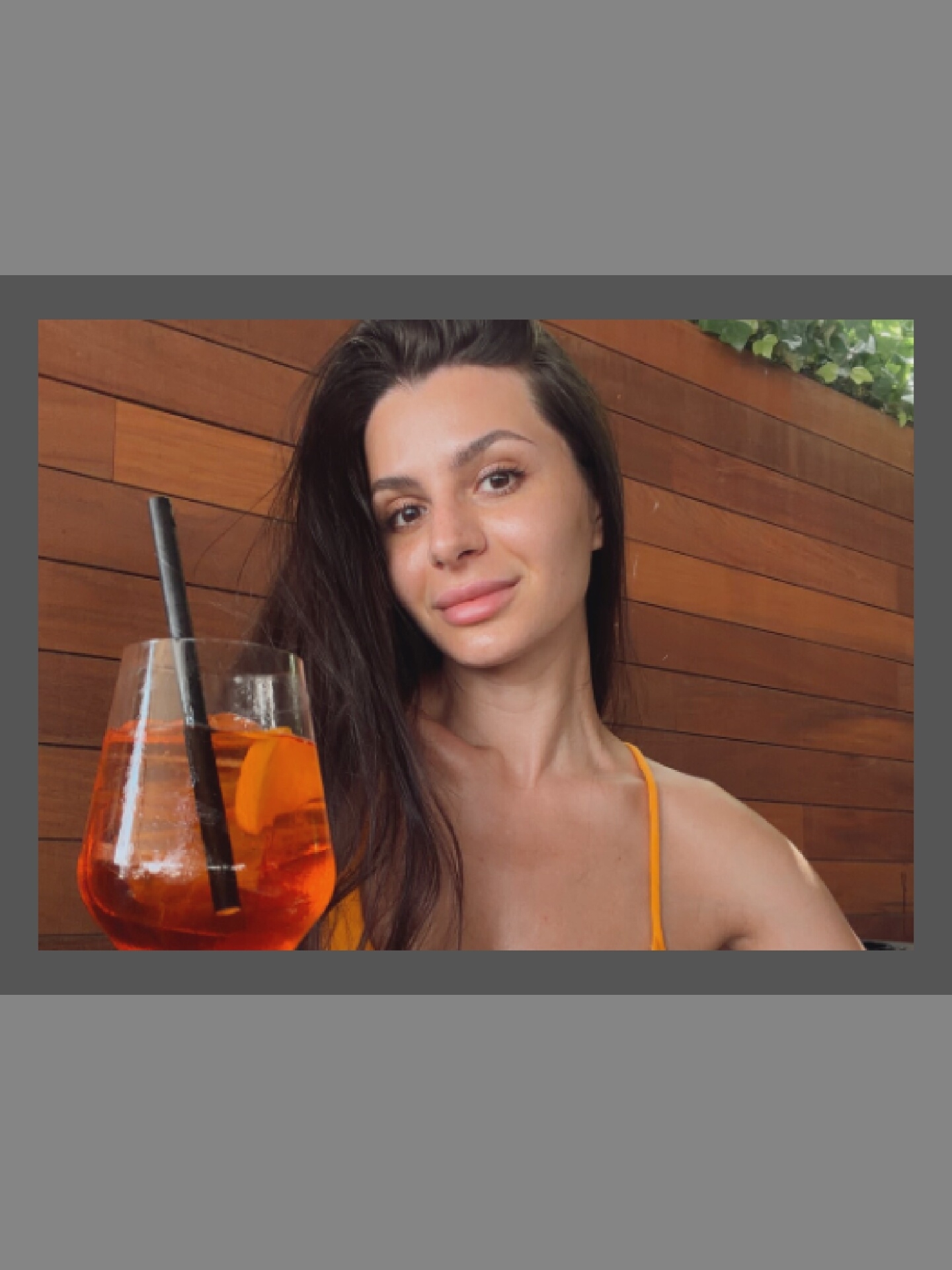 A woman with long dark hair is pictured from the chest up, holding a glass with an orange beverage. The woman is wearing an orange strap top. The background is a wooden wall with some greenery visible at the top. The setting appears to be outdoors.