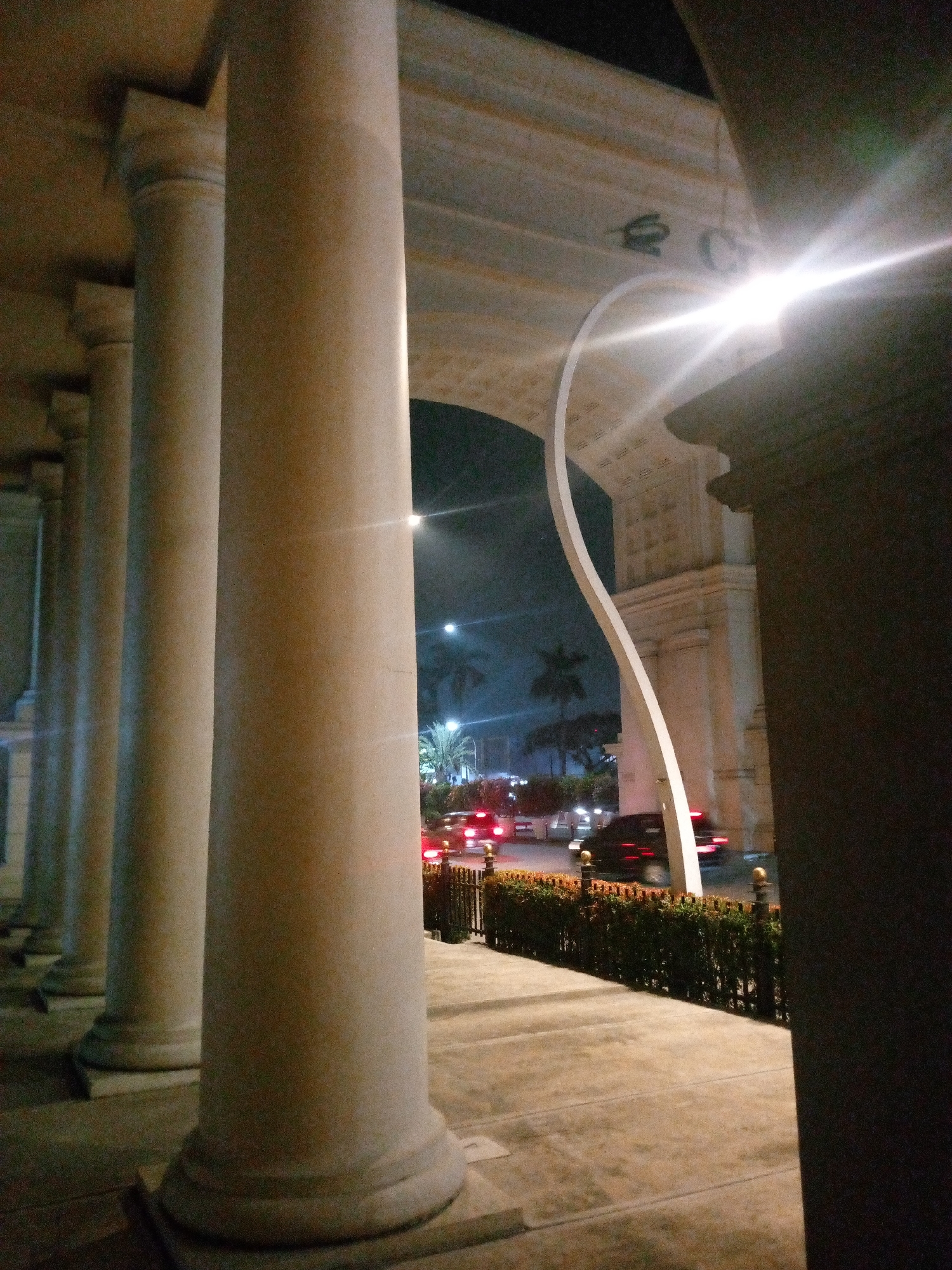 This is a low-light, nighttime photograph taken from a low angle, showing a row of large, smooth, light-colored columns. The columns are positioned in the foreground, with the closest one dominating the right side of the frame. They appear to be part of an architectural structure, possibly a building entrance or a portico, with a curved archway visible in the background.
Beyond the columns, an arched structure is visible against a dark night sky. A bright light source, likely a street lamp or a vehicle's headlights, is causing a significant glare in the upper right quadrant of the image. Through the archway, a street scene is visible. Cars are driving on the road, with their taillights creating streaks of red light, indicating motion. Palm trees and other vegetation are present along the roadside. A low, manicured hedge with decorative posts borders the sidewalk. The ground in the foreground is paved, with visible seams in the concrete or stone. The overall impression is of an illuminated streetscape at night, viewed from a sheltered, colonnaded area.
This photograph is identified as being taken in the city of Bitung Jaya, Indonesia.
