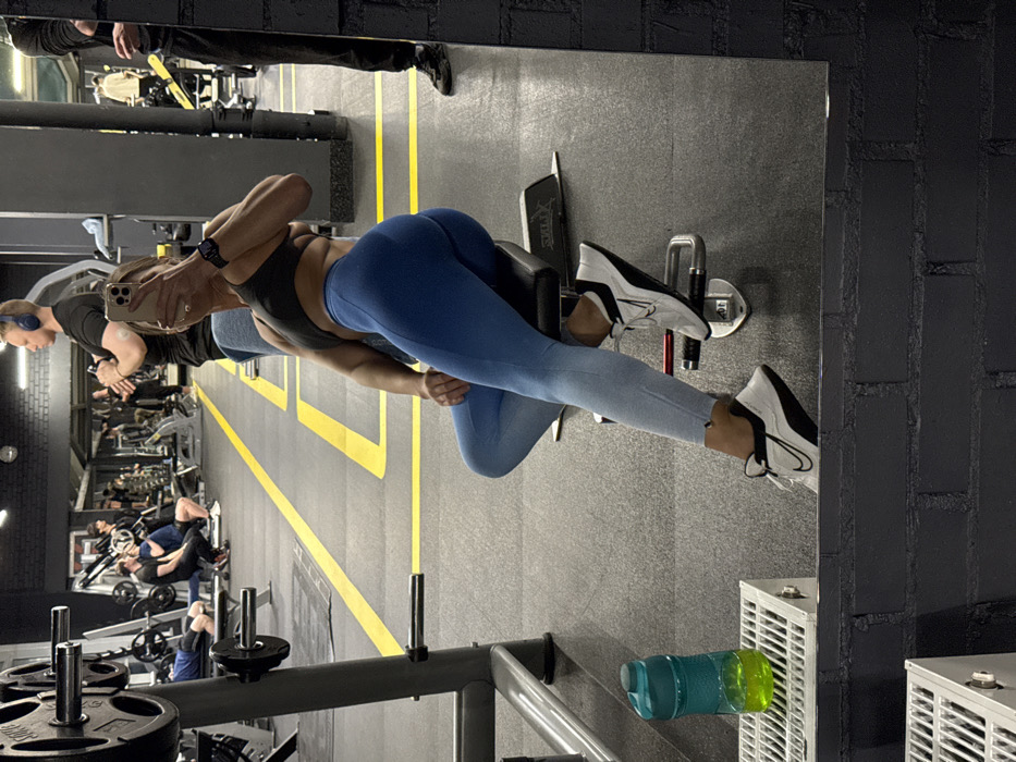 In a gym mirror reflection, a woman in a dark sports bra and blue leggings works out on a leg curl machine. She holds a phone, presumably taking a photo. Behind her, another person uses a similar machine, and other gym patrons are visible in the background. The gym has a gray floor with yellow lines. A water bottle sits near the machine in the foreground.