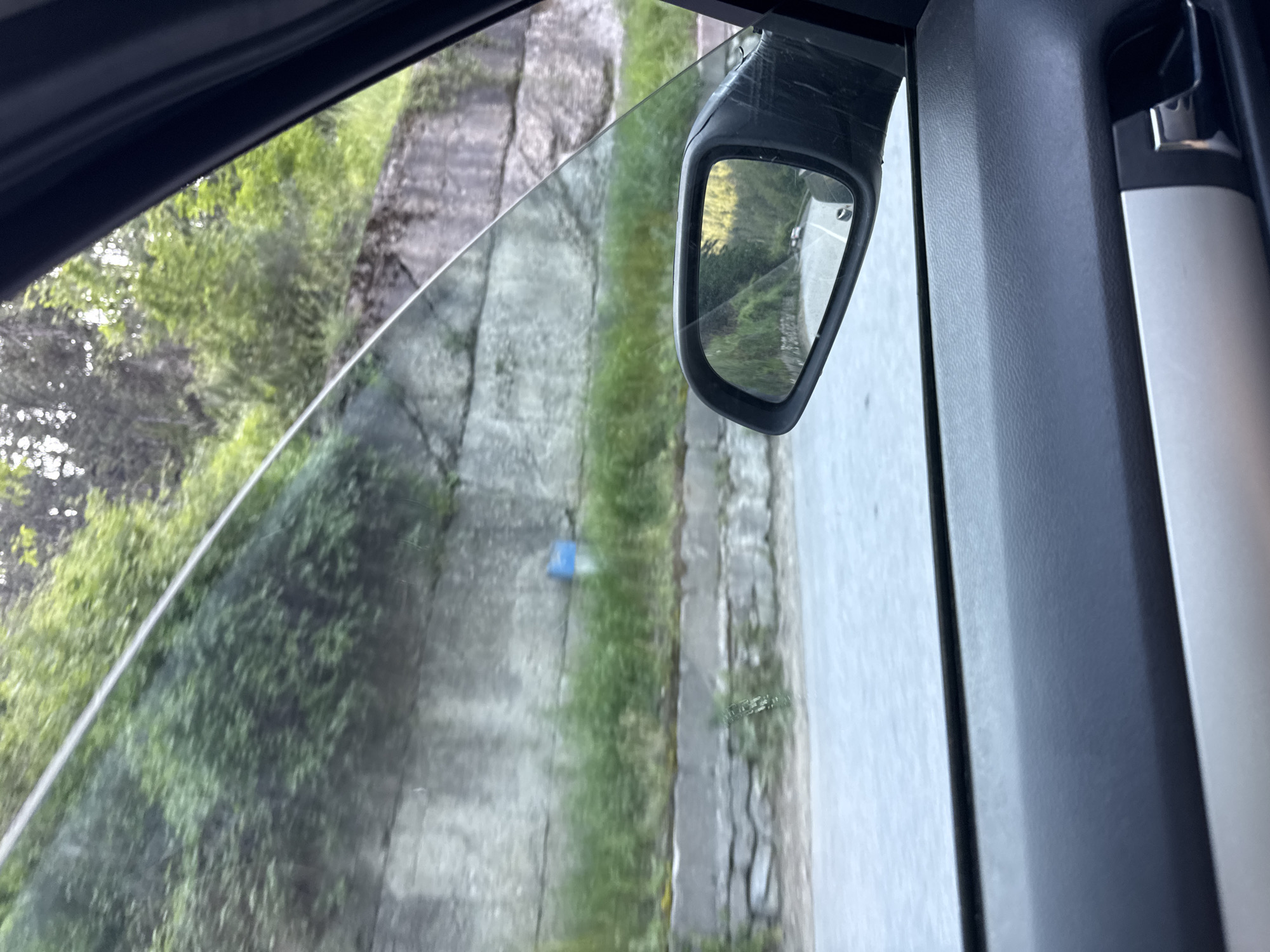 The image depicts a view from the passenger side of a moving vehicle, looking out a window onto a natural and constructed roadside environment, presented as being in Sokolovtsi, Bulgaria. The foreground shows the interior of the car, including a dark textured door panel with a silver-colored door handle and trim at the bottom right. A clear glass windowpane occupies the upper portion of the door, and a black side-view mirror is affixed to the exterior frame, positioned centrally within the window's lower right quadrant.

Through the window, an outdoor scene is visible. A paved asphalt road curves along the lower left. Adjacent to the road, a concrete or stone retaining wall rises, showing signs of wear and some cracking. Green grass and smaller plants grow along the base and intermittently on top of the wall. Further up the embankment, dense green foliage and trees cover a steep slope. A small, partially obscured blue and white marker pole stands against the retaining wall in the midground. The blurred appearance of the vegetation outside the window suggests the vehicle is in motion.

The side-view mirror reflects the road behind the vehicle, showing a winding asphalt road flanked by trees and greenery under bright sunlight. Two distant white vehicles are visible on the road in the mirror's reflection, seemingly moving away from the viewer. No people are explicitly visible in the scene. The overall environment suggests a rural or semi-rural mountainous road.