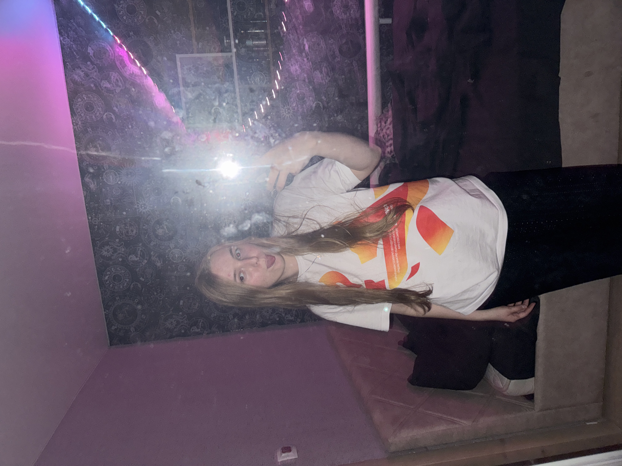 The image depicts a young Caucasian female standing in what appears to be a bedroom, taking a selfie in a mirror. She has long blonde hair and is wearing a white t-shirt with an abstract orange and yellow design and black pants. Her tongue is sticking out. The room has purple walls, and a wall with a celestial-themed wallpaper is visible behind her. LED strip lights are illuminating the room with pink and blue hues, reflecting off the mirror. A bed is partially visible on the left, and a shelf unit with items on it can be seen in the background. The lighting is dim, and the mirror is smudged, obscuring some details. The context provided about the location in the city of Unknown, Russia, is speculative and not directly verifiable from the visual information in the image.