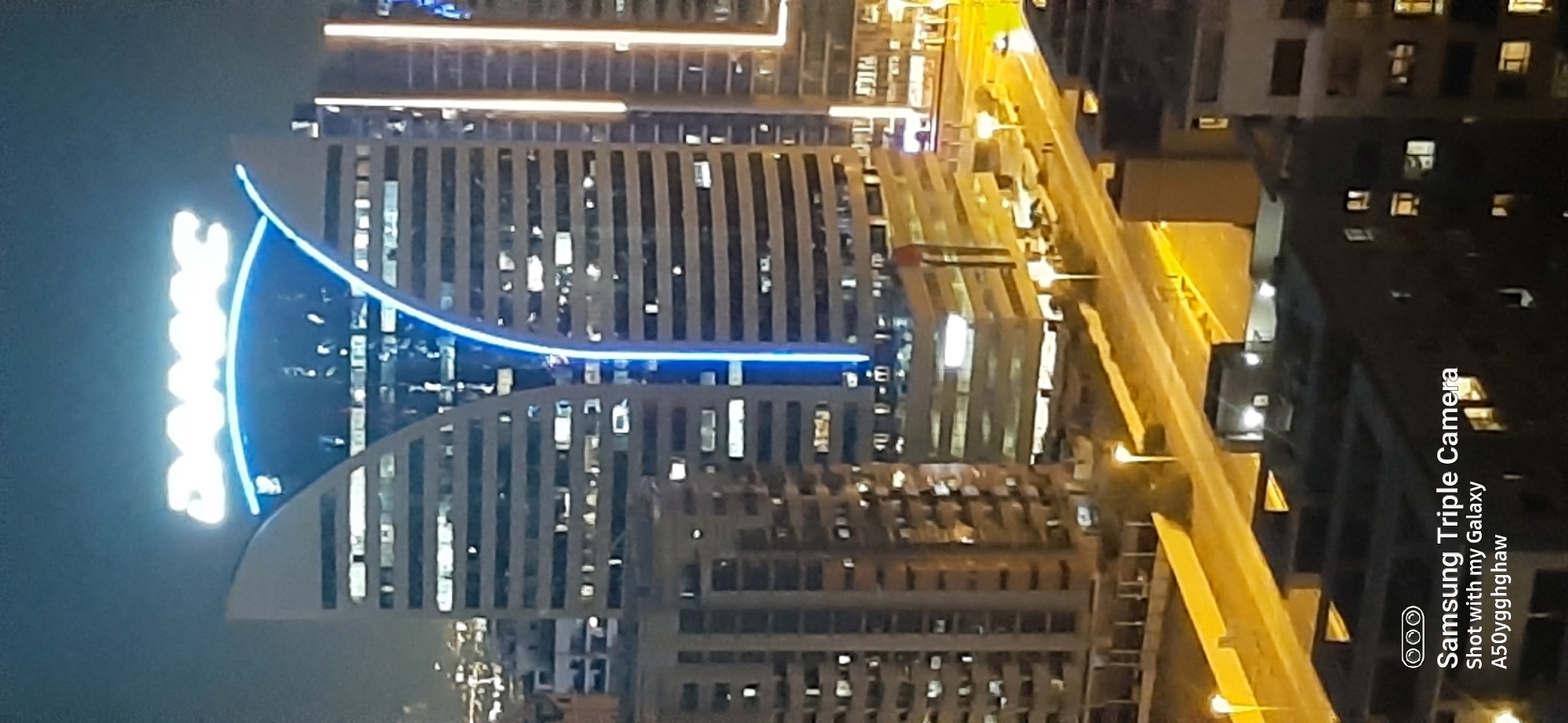 The image captures a nighttime aerial view of modern skyscrapers in Dubai, United Arab Emirates. The prominent feature is a distinctive building with two curved, wing-like structures, illuminated by bright blue neon lights that form a sweeping arc. The building itself has a segmented facade, suggesting multiple floors with lit windows indicating occupancy. Adjacent to this, other high-rise buildings with similar architectural styles and illuminated windows are visible, contributing to a dense urban skyline. A well-lit road with streetlights runs along the base of the buildings, and there is evidence of vehicular traffic. The foreground shows the rooftops of lower buildings. The overall scene depicts a vibrant and illuminated cityscape at night, characteristic of Dubai's modern development.