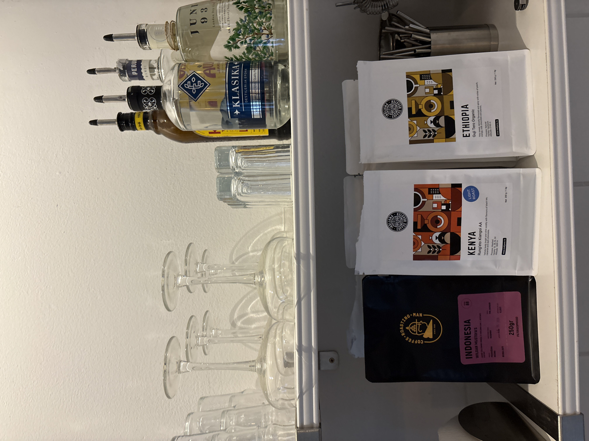 This media file captures a close-up, overhead shot of a home bar or kitchen shelf in Belgrade, Serbia. The subjects are primarily objects: several bottles of spirits with pour spouts, a stack of clear wine glasses, and three bags of coffee. The spirits bottles appear to be different brands, with labels indicating "JUNI 93" and "KLASIKA". The wine glasses are neatly arranged, suggesting they are ready for use. The coffee bags are from "INDJANA COFFEE ROASTERS" and are labeled "ETHIOPIA Guji Tero Organic", "KENYA Rung'eto Kiangoi AA", and "INDONESIA WILDAN MUSTOFA'S". Text on the coffee bags details roast type, flavor profiles, processing methods, and origin. The overall scene conveys a sense of preparation or organization for serving drinks and coffee. The lighting is bright and even, suggesting it could be daytime indoors. There are no people or animals visible.