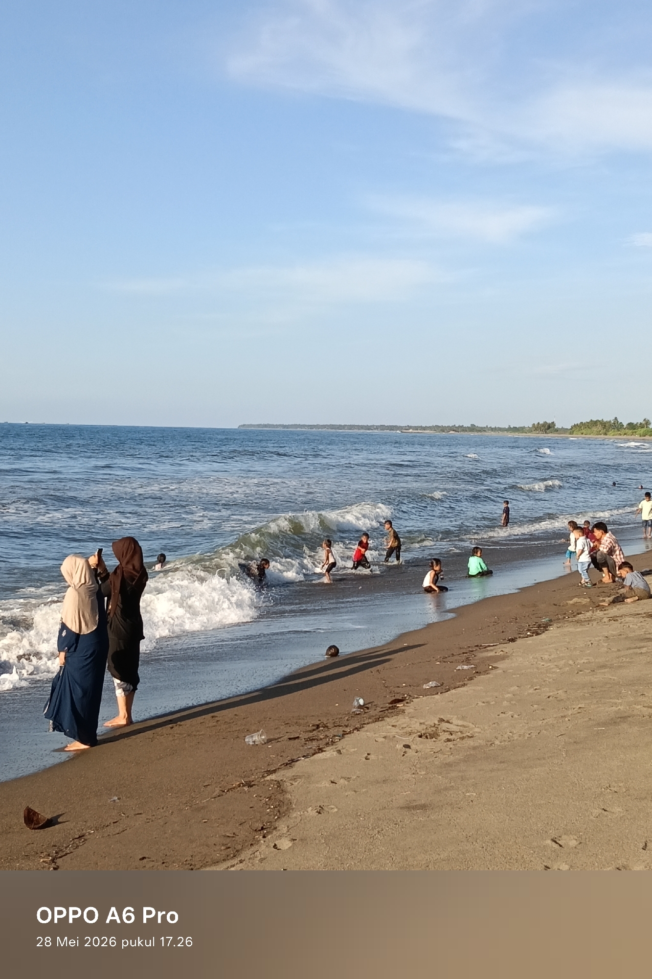The image depicts a beach scene with several people present. Two women, wearing hijabs, stand in the foreground. One is wearing a black hijab and dress and is holding a phone. The other is wearing a cream colored hijab and blue dress. Further down the shoreline, a group of children and adults are playing and wading in the water. The beach is sandy and the water is dark blue with white waves. In the distance, the coastline is visible with some trees. There is a clear blue sky with some wispy clouds. The location is Rheumbaroh, Indonesia.
