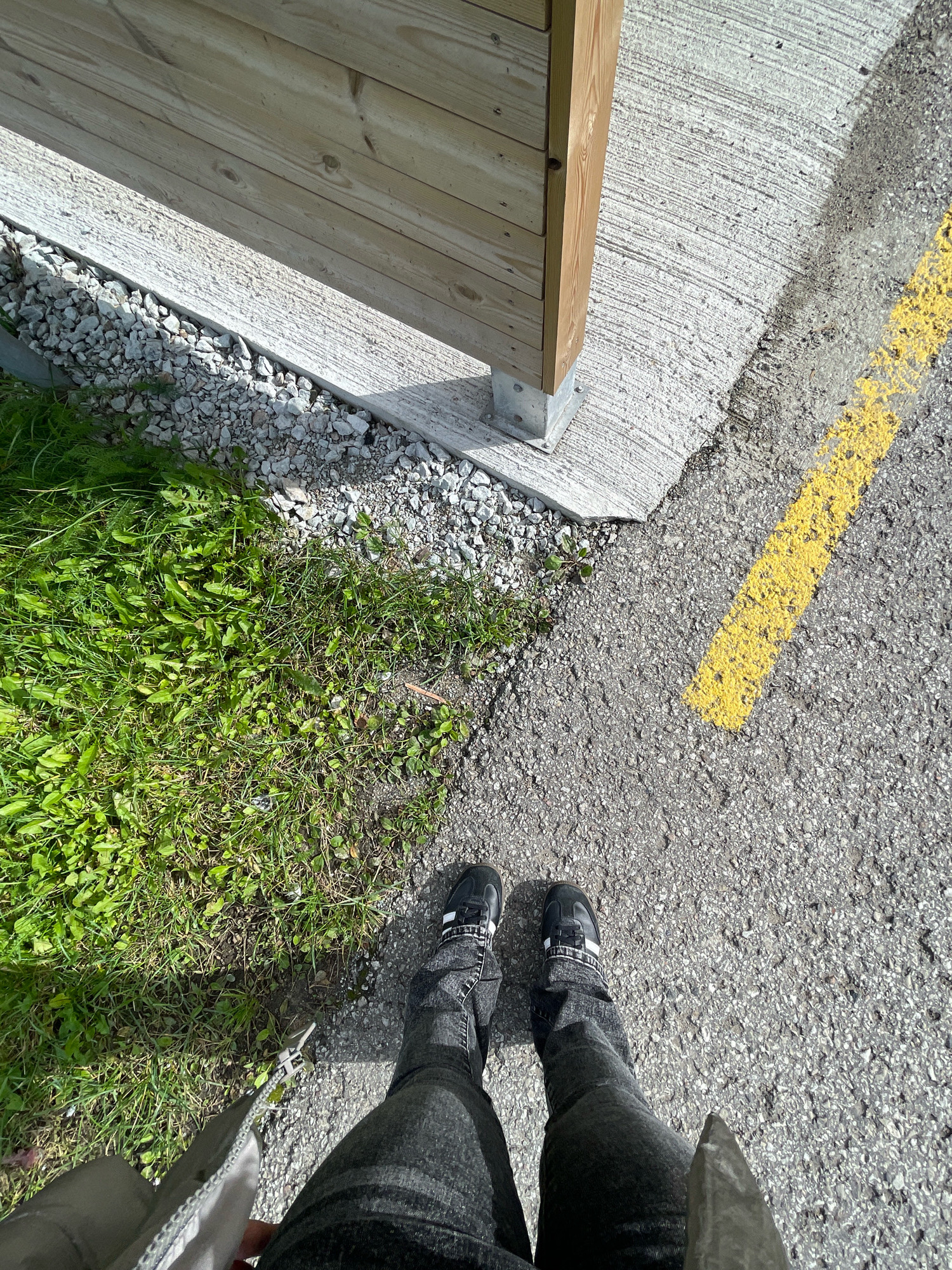 A high-angle, full-body shot shows a person standing in a paved area next to a grassy patch and a wooden structure. The person, wearing dark pants and dark sneakers with white accents, is facing away from the camera. The ground is a mix of asphalt and concrete, with a bright yellow line painted on the asphalt. To the left, there is a patch of green grass and a border of small gray rocks. Behind the rocks is a wooden fence or wall made of horizontal planks, supported by a metal post. The lighting suggests a sunny day with shadows cast on the ground and the wooden structure. The scene appears to be an outdoor public space, possibly a park or a walkway. There is no indication of the specific location in Tallinn, Estonia.