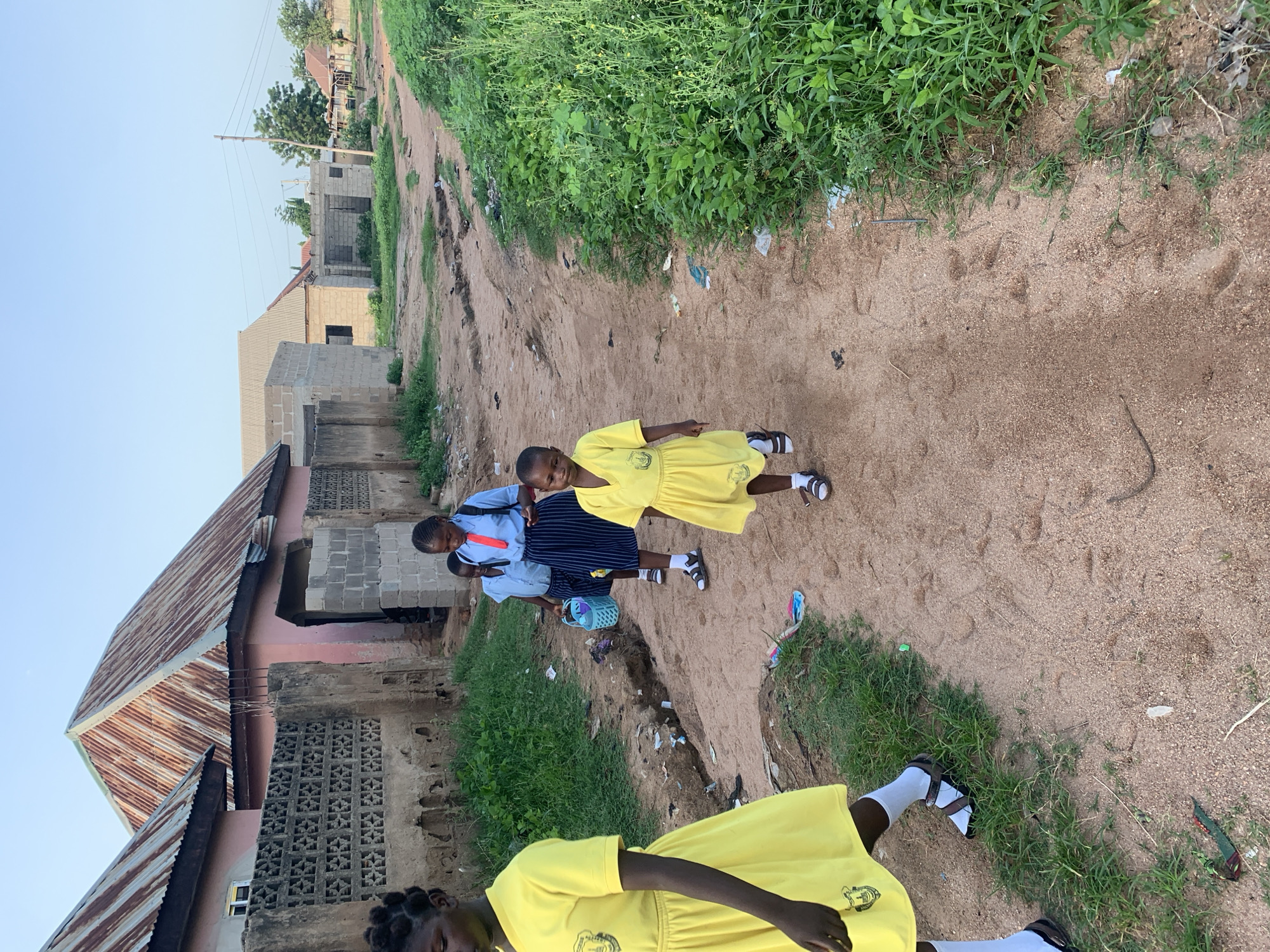 The image depicts a dirt road in Jalingo, Nigeria, with several children walking on it. The children are dressed in school uniforms, primarily yellow dresses with emblems, and one child wears a blue shirt and a striped skirt with a red tie. They appear to be walking in the same direction. The environment is characterized by a dusty, uneven road, bordered by vegetation and residential buildings. Some of the buildings have corrugated metal roofs, while others are constructed from concrete blocks. There are also remnants of trash and debris scattered along the roadside. The sky is clear and light blue, suggesting it is daytime. The overall scene conveys a sense of everyday life in a Nigerian community, with children on their way to or from school.