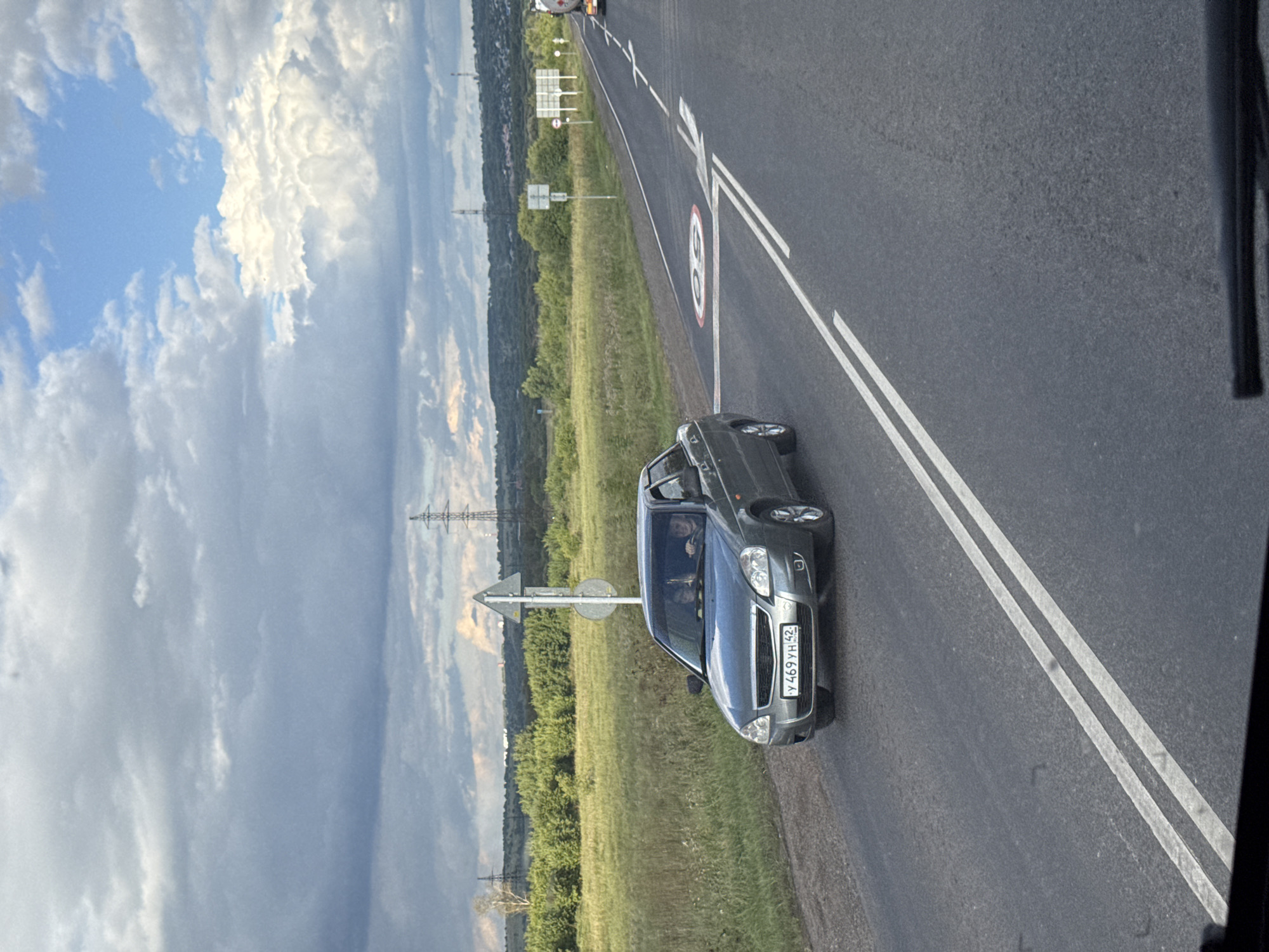 A gray sedan, identified by license plate Y 469 YH 42, is traveling on a two-lane asphalt road under a cloudy sky. The car is in the left lane, with two occupants visible in the front seats. A speed limit sign indicating 50 is painted on the road. The road is lined with white double lines. To the left of the road, a grassy field with scattered trees stretches towards a line of hills in the distance, where power lines are visible. A traffic sign is present on the left side of the road. In the background, a tanker truck is visible on the right lane of the road, moving away from the camera.