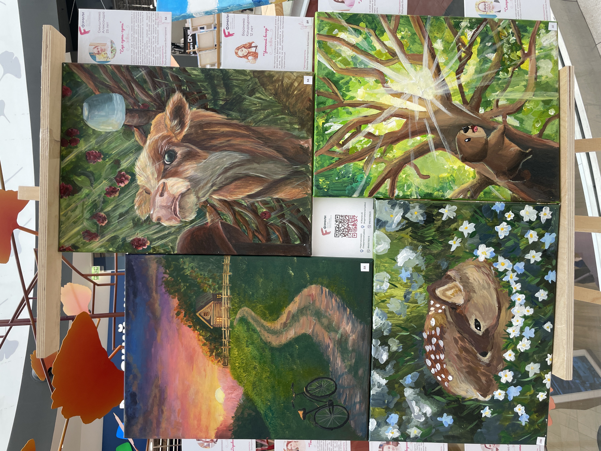 This media file features a collection of four paintings displayed on easels. The overall scene appears to be an art exhibition or a display of student artwork, possibly in Perm, Russia, as indicated by some visible text.
The top-left painting is a close-up of a cow-like animal with large eyes, set against a background of green foliage and red berries. Above it, a small, light blue, reflective object hangs from a branch. The top-right painting depicts a tree with sunlight streaming through its branches, creating a radiant effect. A small, brown creature with its tongue sticking out is shown hanging from a branch.
The bottom-left painting is a landscape scene at sunset. A winding dirt path leads towards a small wooden house with illuminated windows, nestled amongst rolling green hills. A bicycle is parked near the path. The sky transitions from deep blue to vibrant pink and orange hues.
The bottom-right painting is a close-up of a young deer curled up amidst white and light blue flowers. The deer has a spotted coat and appears to be sleeping or resting peacefully.
Visible text includes artist names and ages, such as "Смышляева Александра 26 лет" (Smyshlyaeva Alexandra, 26 years old) and "Вероника Кондаурова 12 лет" (Veronika Kondaurova, 12 years old), along with titles for the paintings like "Еще чуть-чуть" (A little bit more) and "Уютный вечер" (Cozy evening). There are also indications of instructors like "Зайцева Виктория Викторовна" (Zaitseva Victoria Viktorovna) and "Белова Екатерина Викторовна" (Belova Ekaterina Viktorovna), and addresses such as "Крестьянская, 6" and "В.Засулич 42", along with phone numbers. The overall mood conveyed by the paintings is one of nature, tranquility, and youthful artistic expression.