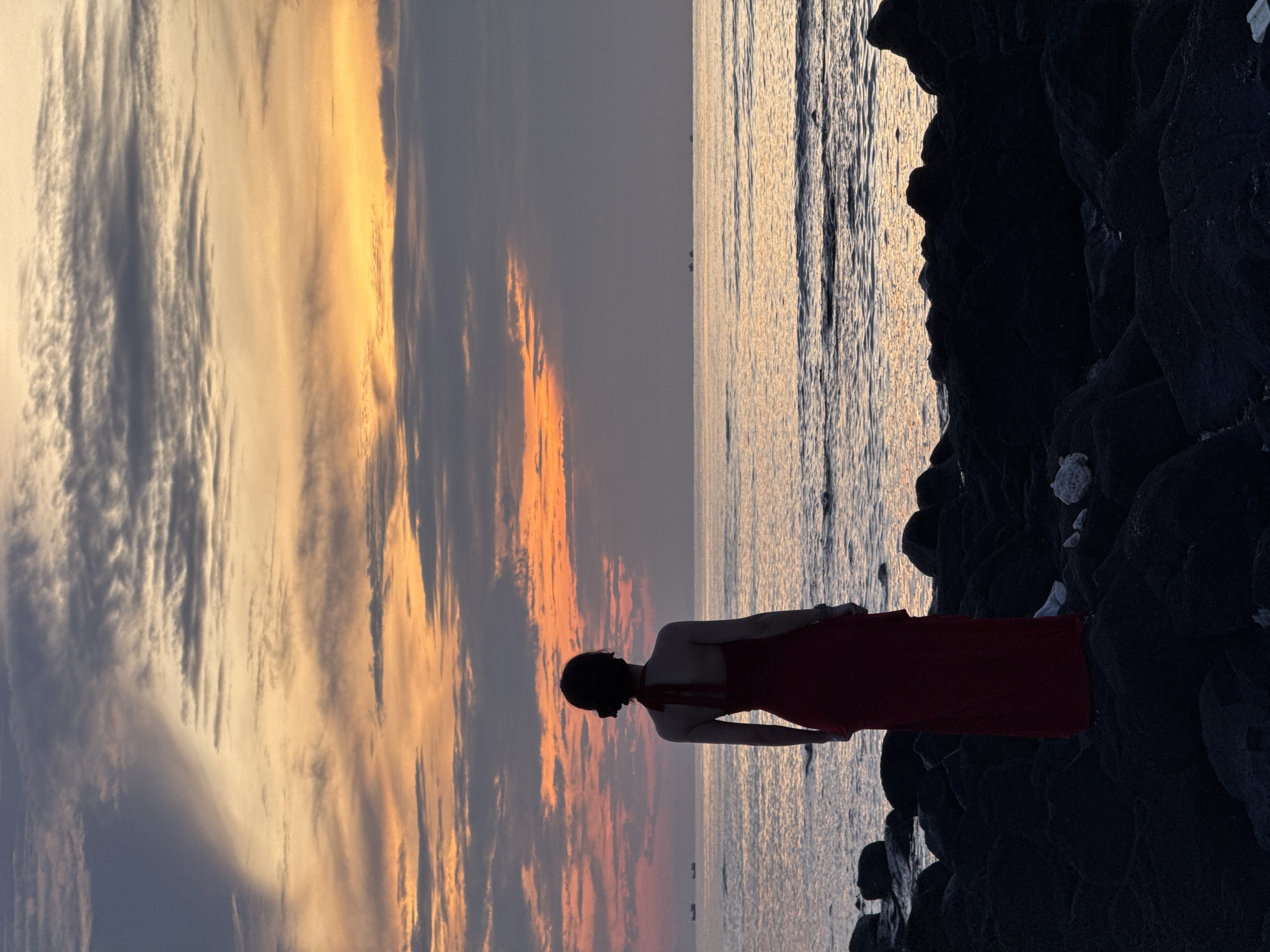 A lone woman in a flowing red dress stands on a rocky shore, facing a serene ocean at sunset. The sky is a canvas of warm colors, with hues of orange, yellow, and purple blending into soft gray clouds. The gentle waves of the sea reflect the fading light, creating a tranquil and picturesque scene. In the distance, a few small boats can be seen on the horizon. The woman's silhouette, against the dramatic sky and sea, adds a touch of human presence to the vastness of nature. The overall atmosphere is one of peaceful contemplation and natural beauty. This scene is characteristic of the coastal landscapes of Lý Sơn Special Administrative Region, Vietnam, known for its volcanic rock formations and stunning sunsets.