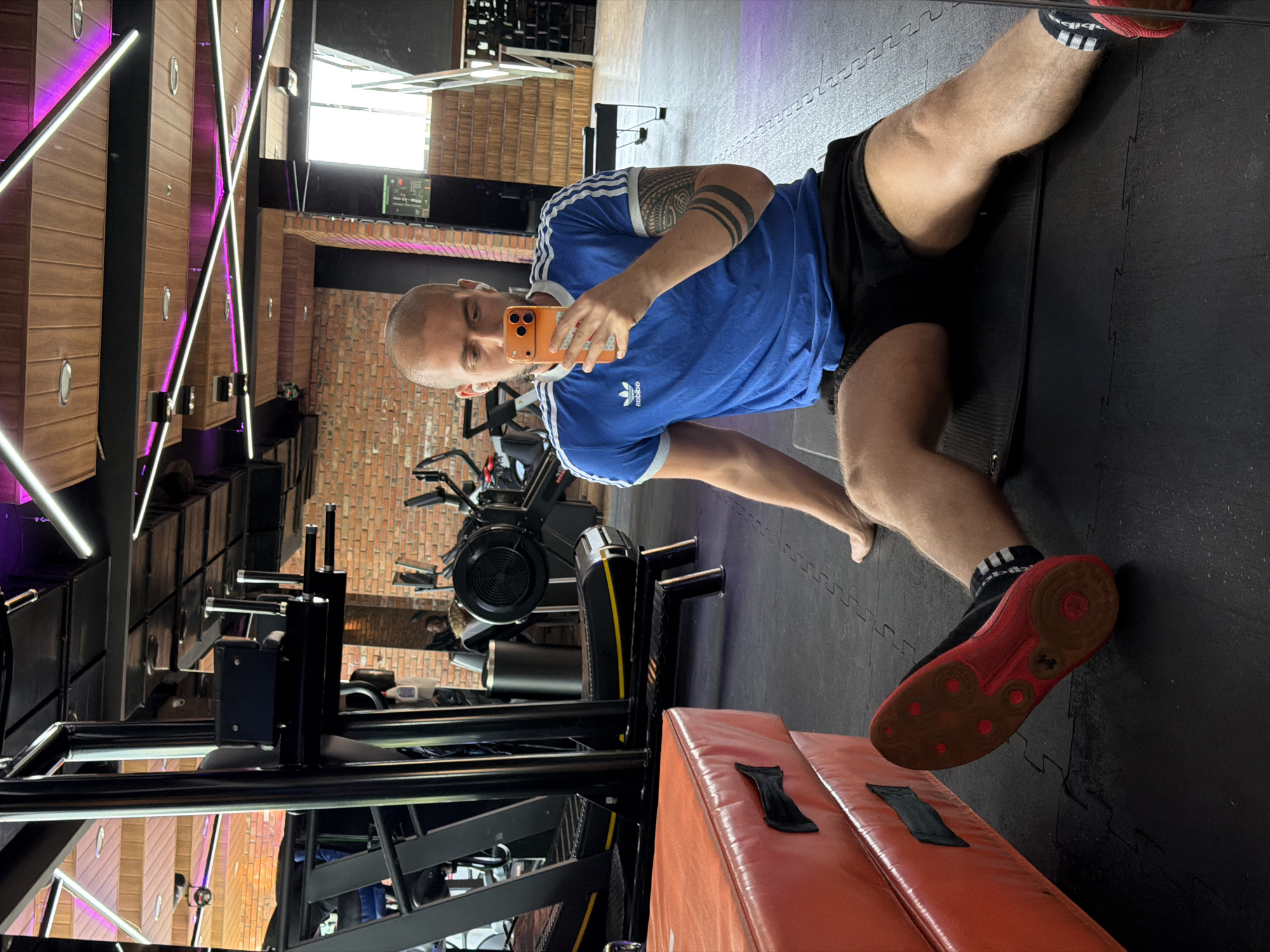 This media file captures a full-body selfie taken by a bald, muscular man in a gym. He is wearing a blue Adidas t-shirt with white stripes and black athletic shorts. The man has a tattoo on his left arm and is holding an orange smartphone to take the photo. He is lying on a black exercise mat with his legs extended. His feet are clad in red athletic shoes with black accents and white Adidas socks.
The gym setting is evident from the exercise equipment in the background, including rowing machines and other weight training apparatus. The walls appear to be brick, and there are modern lighting fixtures with purple and white lights, suggesting a contemporary gym environment. The floor is covered in black interlocking mats.
The man's posture and the gym setting suggest he is engaged in a workout or has just completed one. His expression is neutral, and the overall mood of the image is one of fitness and activity. The presence of gym equipment and the professional lighting indicate this is likely a commercial gym. The time of day is not explicitly clear, but the artificial lighting suggests it could be evening or a dimly lit indoor space. The location is Sofia, Bulgaria. There is no visible text in the image other than the Adidas logo on the man's shirt and possibly on his socks.