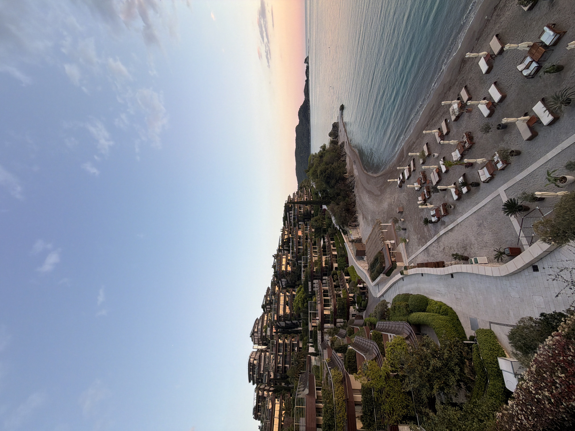 The image captures a tranquil coastal scene, identified as Budva, Montenegro, viewed from an elevated vantage point during what appears to be either sunrise or sunset. The light conditions create a soft, serene atmosphere.
The sky dominates the upper portion of the frame, displaying a clear, light blue hue with sparse, wispy clouds. Towards the horizon, the sky transitions into delicate shades of pink and orange, indicative of either dawn or dusk.
Below the sky, the calm, light blue-green sea extends to meet a wide, curving beach. The water shows gentle ripples, reflecting the soft light. A small, rocky headland or pier juts into the water from the beach.
The beach itself is lined with numerous neatly arranged, empty beach loungers and sunbeds, typically found at a resort. These loungers feature white cushions and coverings, and some are accompanied by potted plants.
Along the hillside above the beach, a modern, terraced architectural complex is integrated into the landscape. These multi-story buildings, likely part of a luxury resort or residential area, feature numerous windows and balconies, and are surrounded by lush, manicured greenery, including hedges, trees, and shrubs. Winding, paved pathways and meticulously landscaped garden areas lead down through the complex towards the beach. The design of the buildings seems to blend seamlessly with the natural contours of the hill.
No people are visible on the beach or within the resort area, lending a sense of peace and quiet to the scene. There is no specific activity taking place, emphasizing the tranquil nature of the moment. No text is visible in the image.